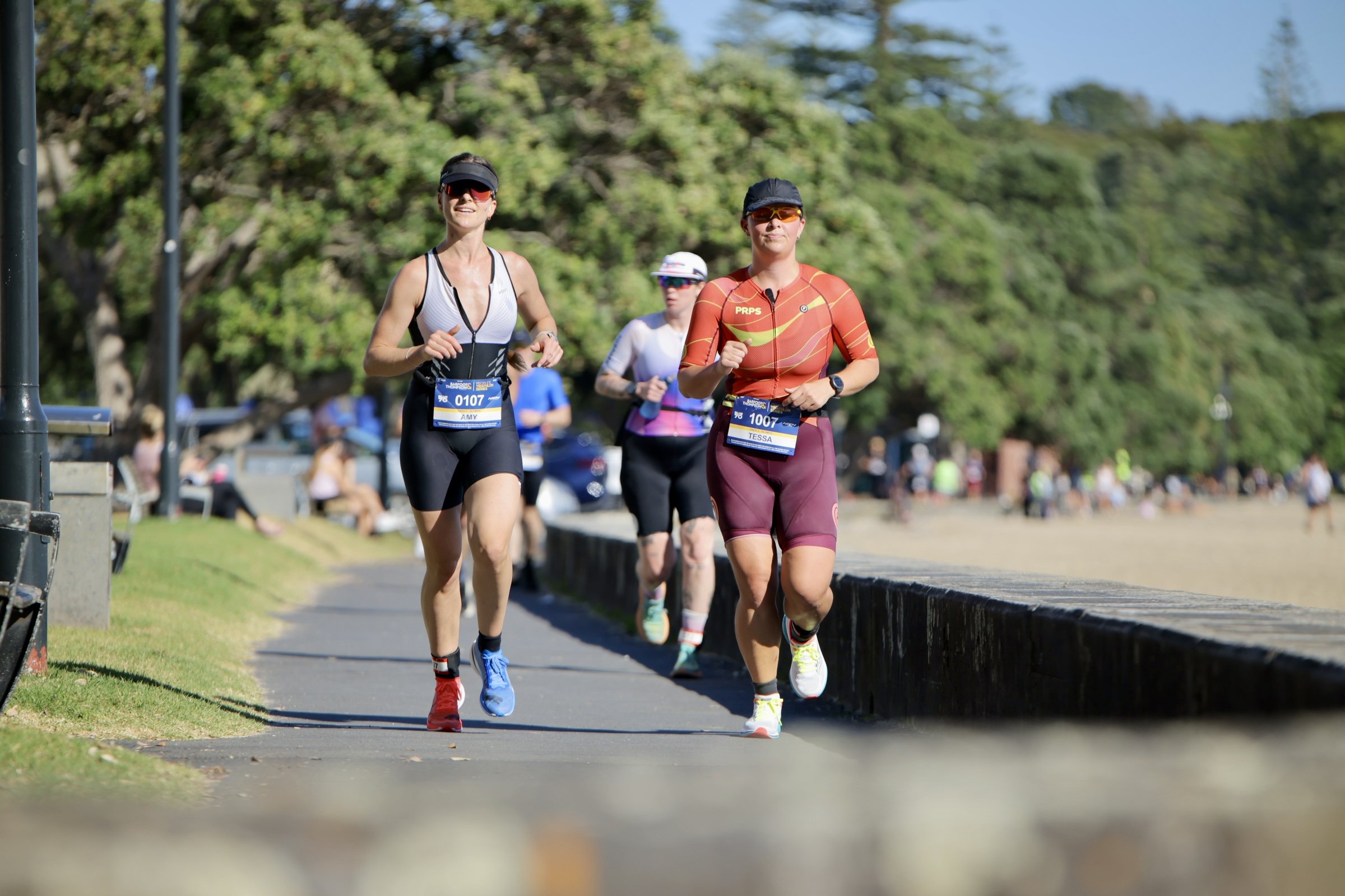 Two women running in a race or marathon on a paved pathway lined with grass, with trees in the background. Both women are wearing athletic gear and sunglasses, and have race bibs with their names and numbers.