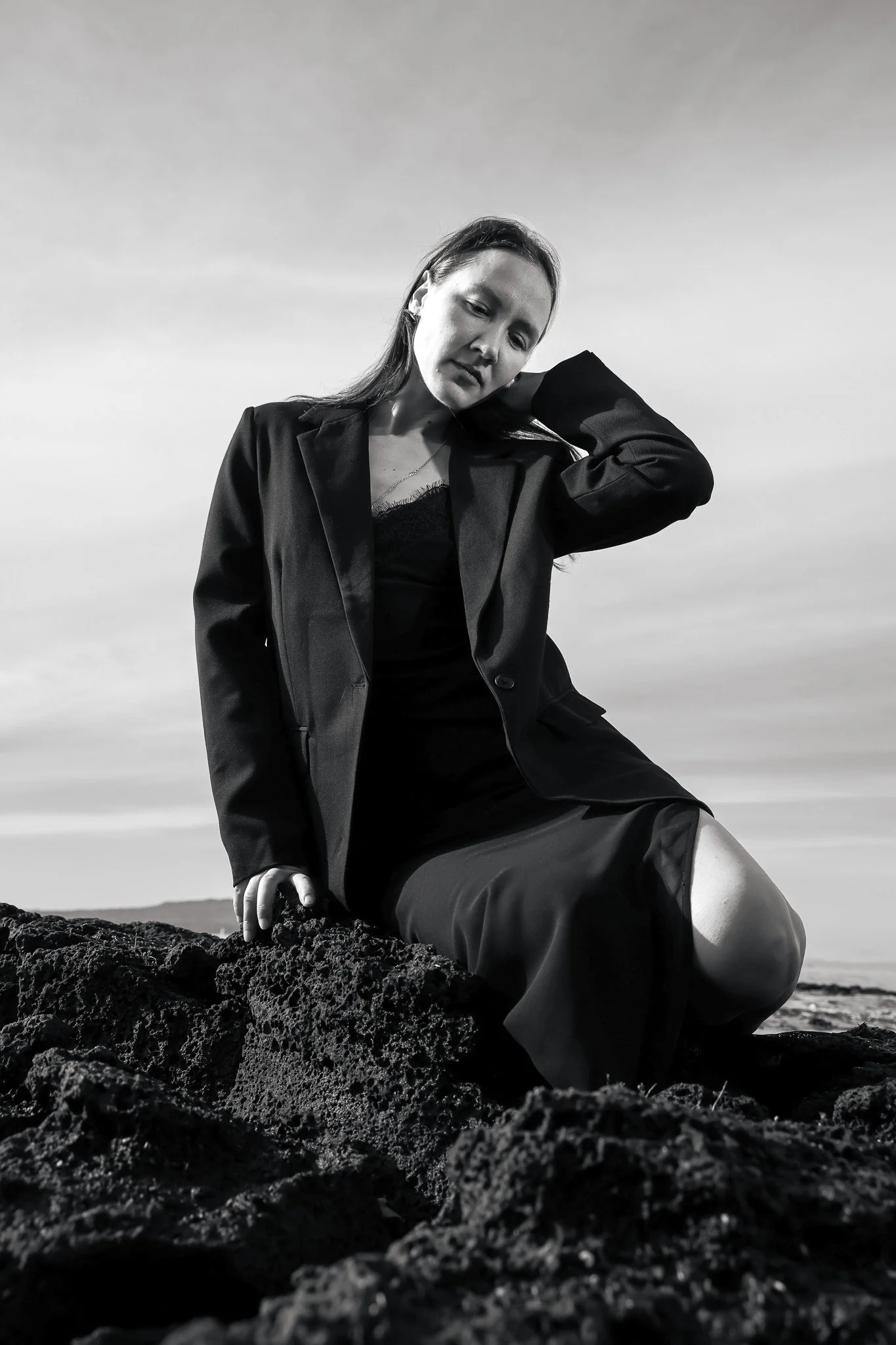 A woman with long hair sitting on volcanic rocks at the beach, dressed in a blazer and shorts, looking contemplative with her hand on her neck, in black and white.