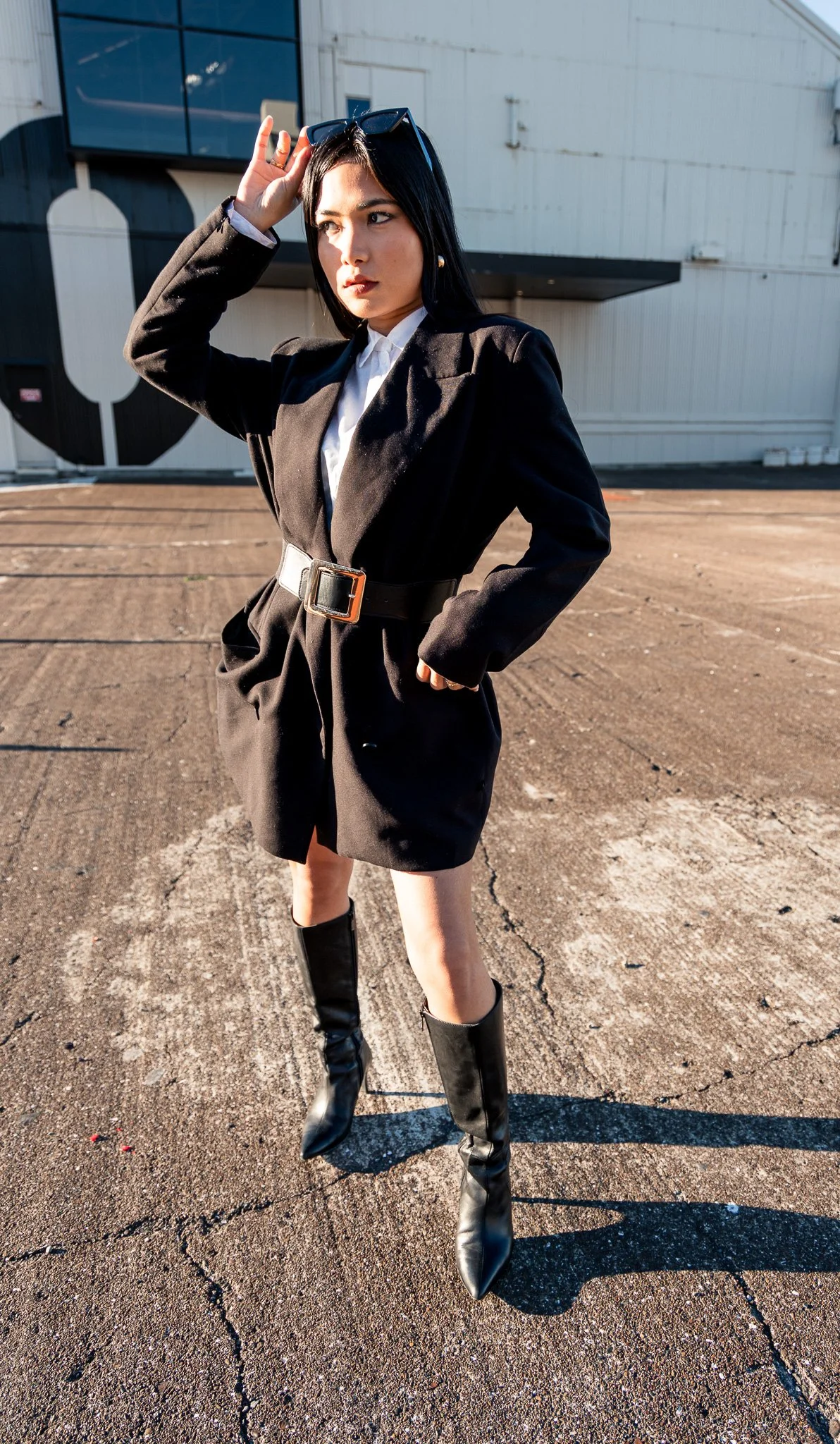 A woman with long black hair and sunglasses on her head standing outdoors on a cracked asphalt surface in front of a white industrial building, dressed in a black blazer belted at the waist, black shorts, and tall black boots, with sunlight casting s