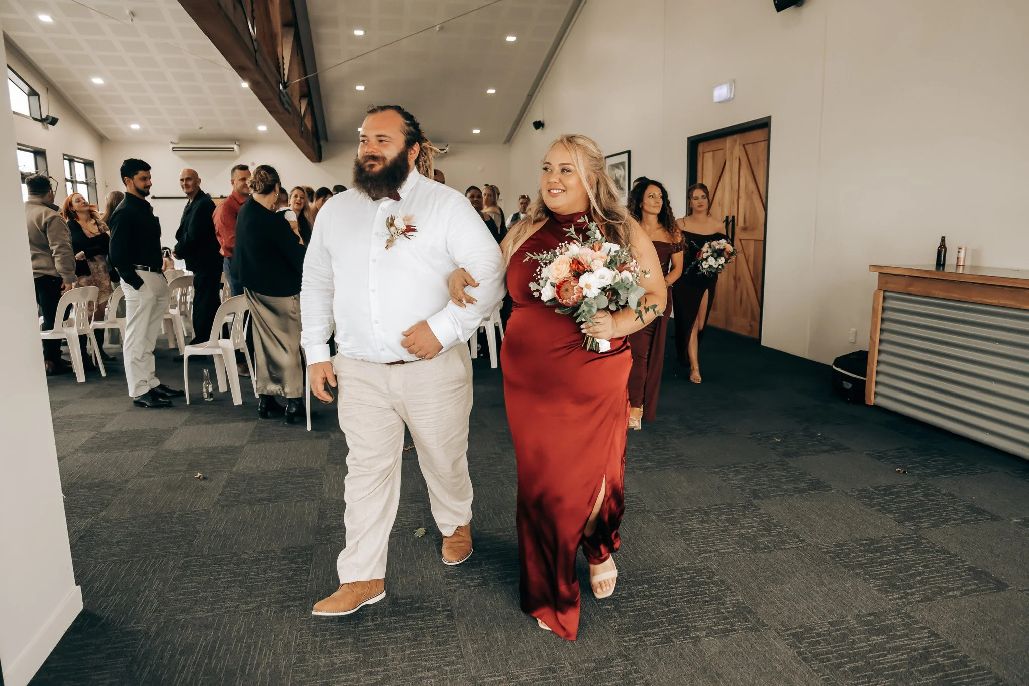 A woman in a red dress holding a bouquet of flowers walking arm-in-arm with a man in a white shirt and beige pants at a wedding reception. Guests are standing and sitting in the background, in a decorated indoor venue.