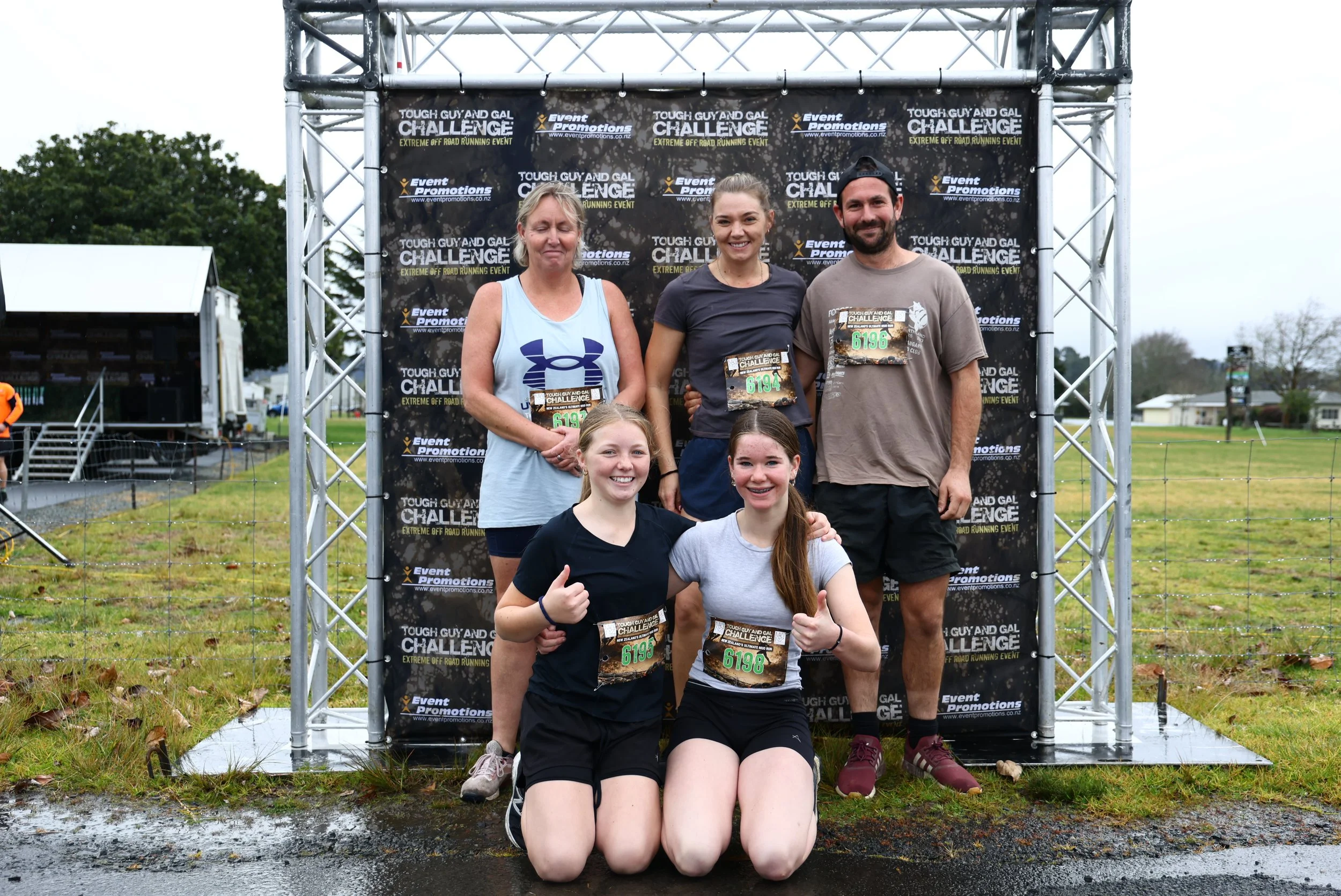 Group of six people, four women and two men, posing in front of a backdrop at an outdoor event. They are participating in an extreme off-road running event called 'Tough Guy and Gal Challenge.' Some people are kneeling in front, others standing behin