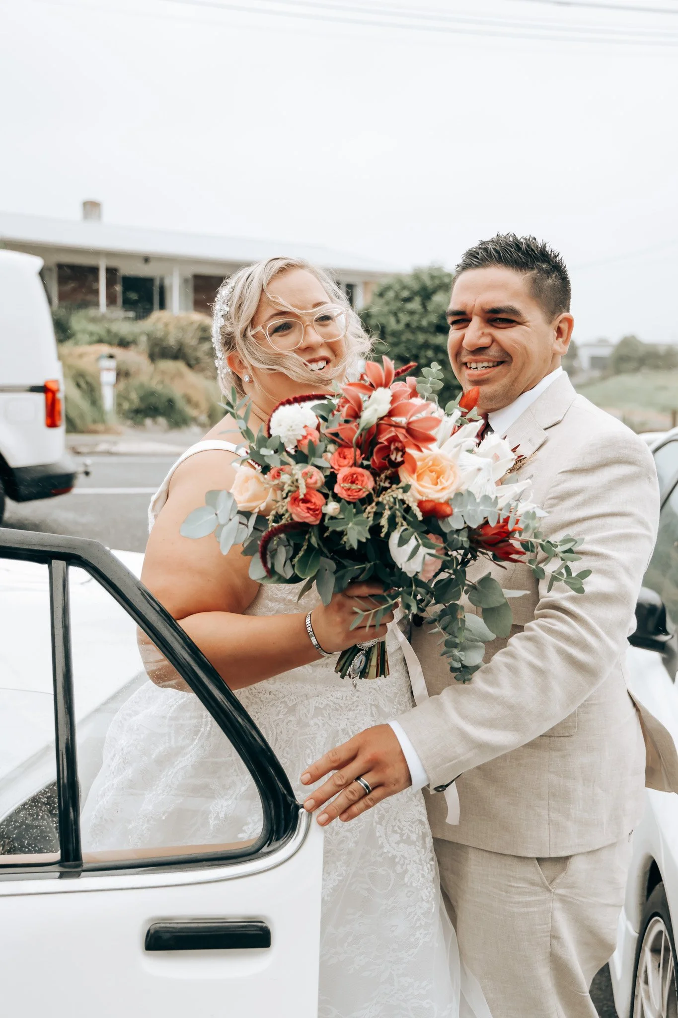 A bride and groom smiling and holding a bouquet of flowers in front of a white vintage car, outdoors with a house and greenery in the background.