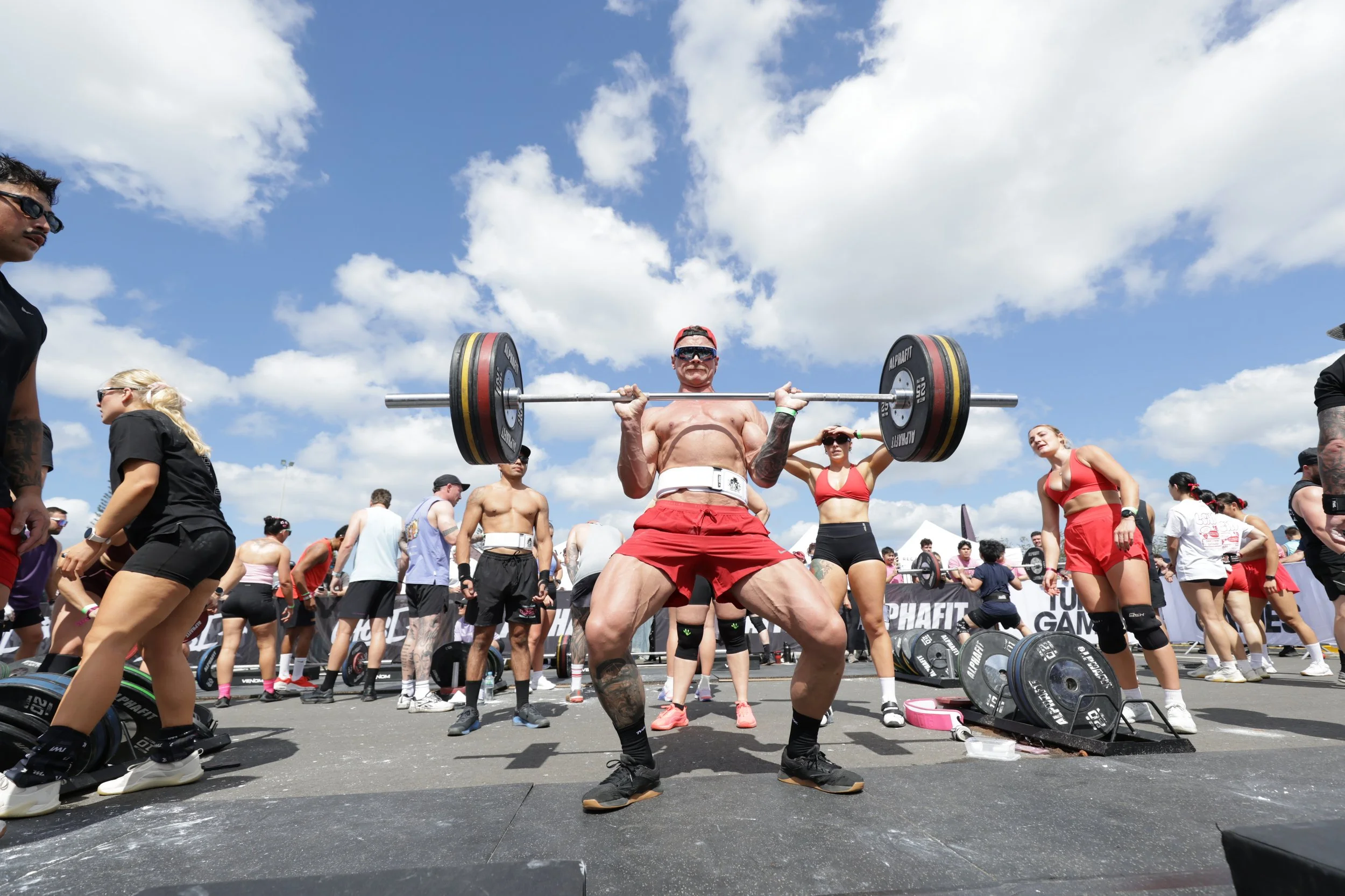 A muscular man wearing sunglasses and a red cap lifts a weighted barbell at an outdoor fitness event, surrounded by a crowd of people, some in athletic clothing, under a partly cloudy sky.