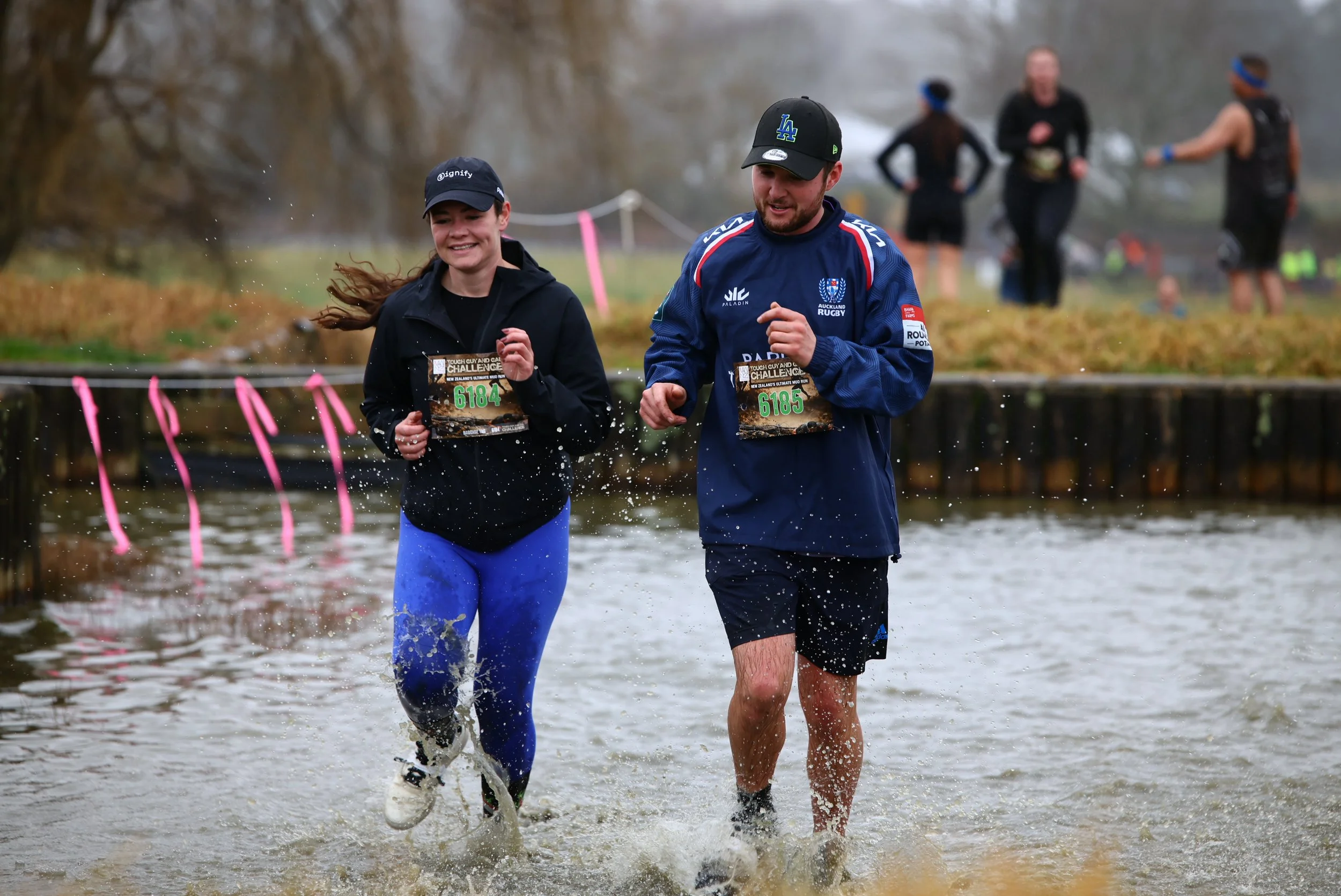 Two people running through water outdoors during a race, with others in the background. The person on the left is wearing a navy cap and a black hoodie, smiling. The person on the right is wearing a black cap, blue sports jacket with logo, and black 