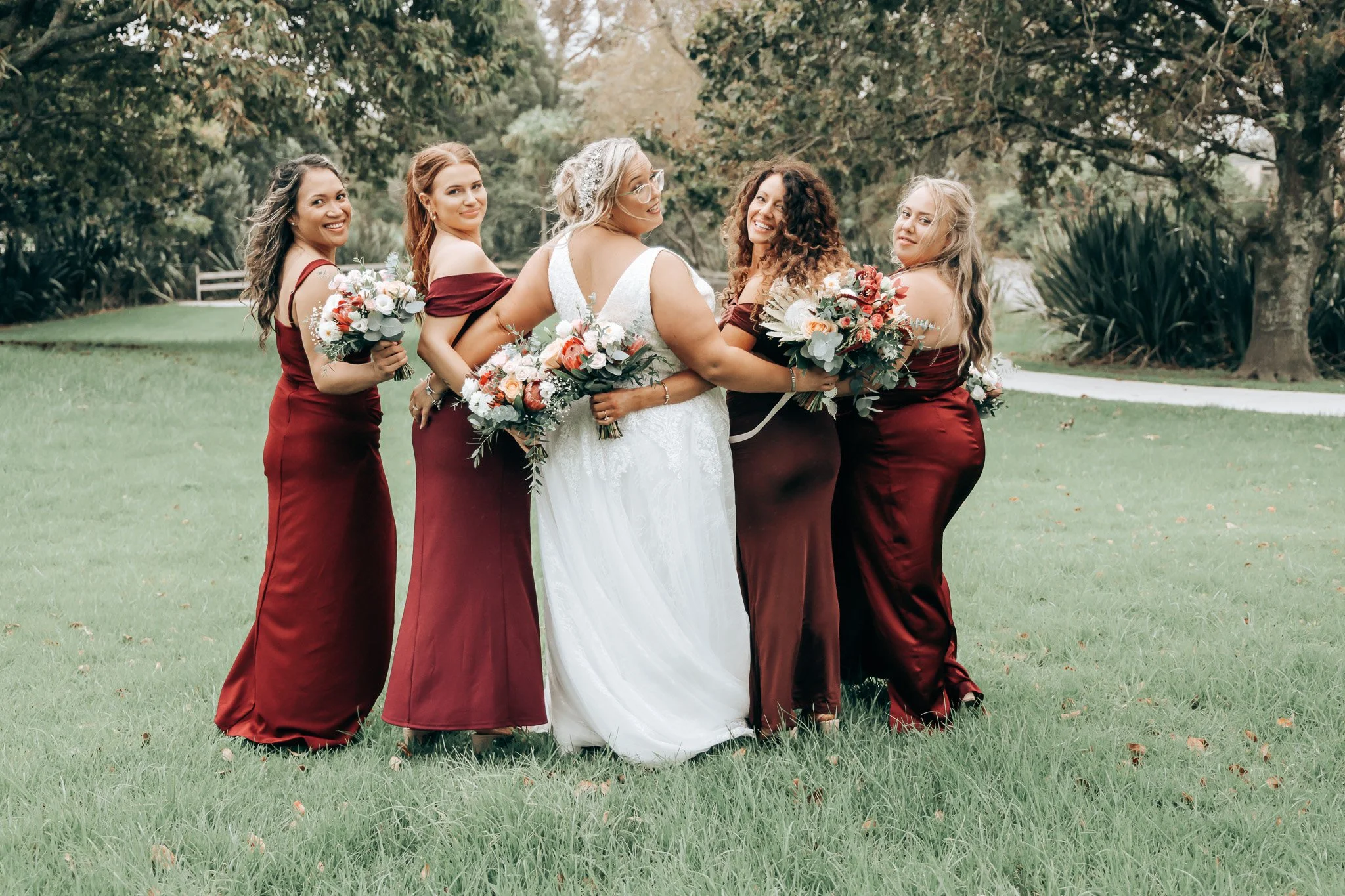 A bride in a white wedding dress surrounded by five bridesmaids in burgundy dresses, all holding bouquets, outdoors in a park with trees and grass.
