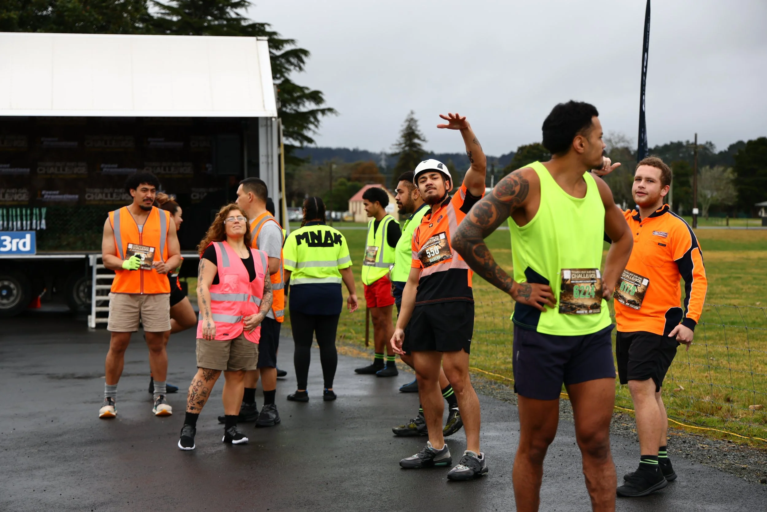 A group of people participating in an outdoor race or marathon event, standing on a paved surface with some wearing race bibs and bright clothing, and others in safety vests. There is a truck in the background and trees under an overcast sky.