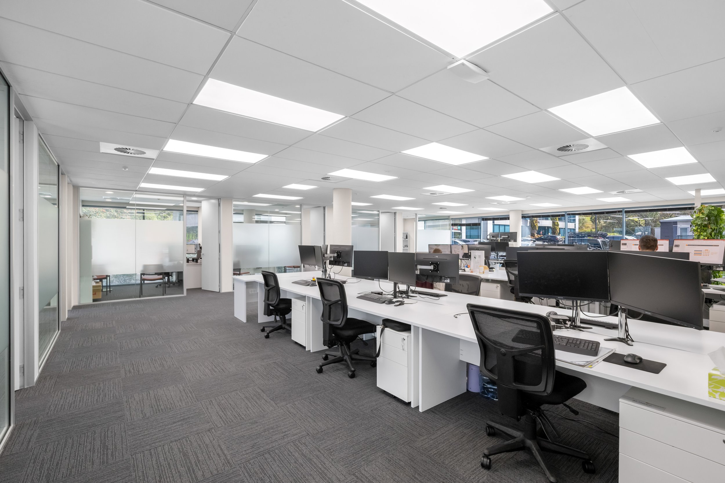 Modern office space with white desks, multiple computer monitors, black office chairs, large windows, and overhead fluorescent lighting.
