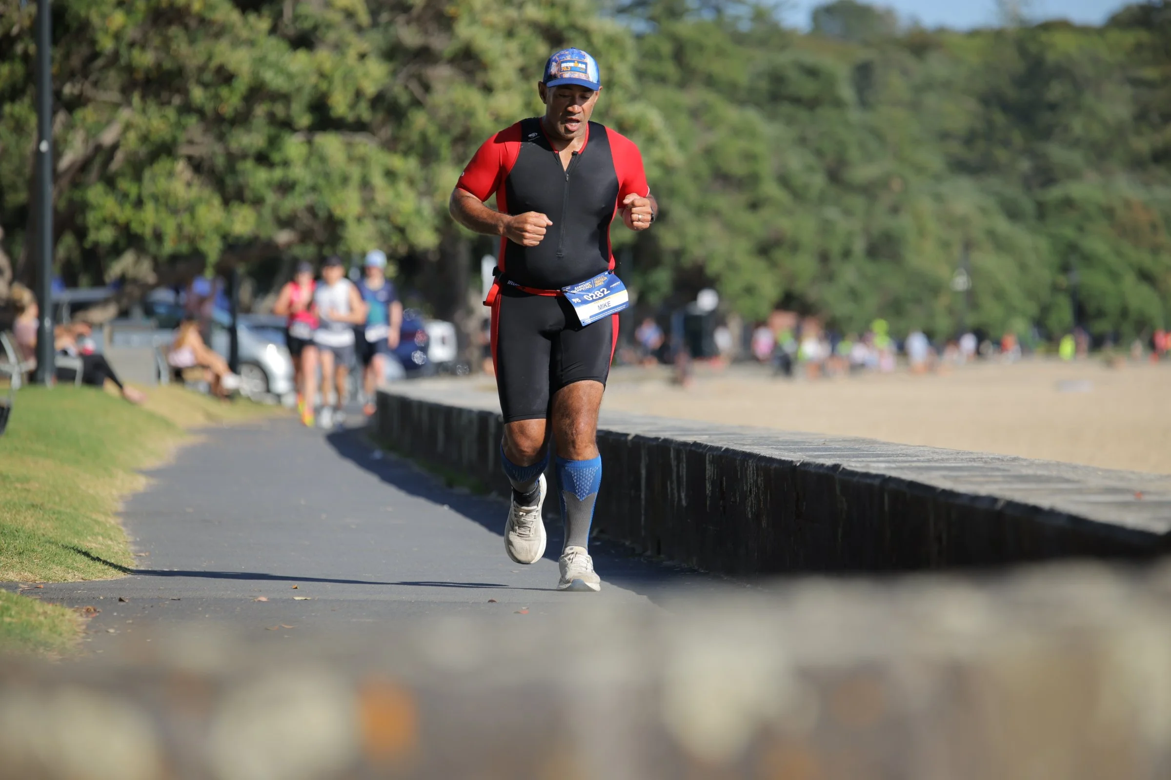A man in athletic gear running outdoors on a sidewalk during a race, with a group of runners behind him and trees in the background.