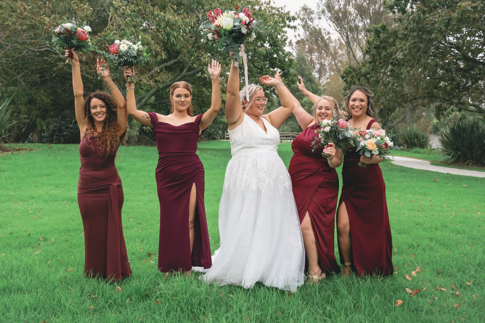 A group of five women celebrating outdoors on a grassy area with trees in the background. One woman is wearing a white wedding dress and tiara, while the other four are wearing matching burgundy dresses. The women are smiling and holding bouquets of 