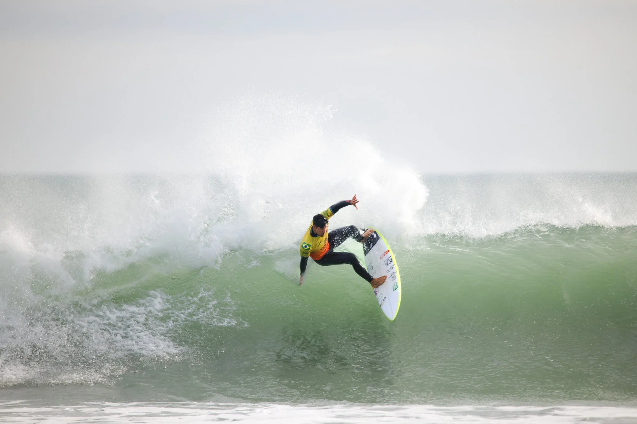 A male surfer in yellow and black wetsuit riding a wave on a white surfboard with a green fin, splashing water as he maneuvers on the ocean.