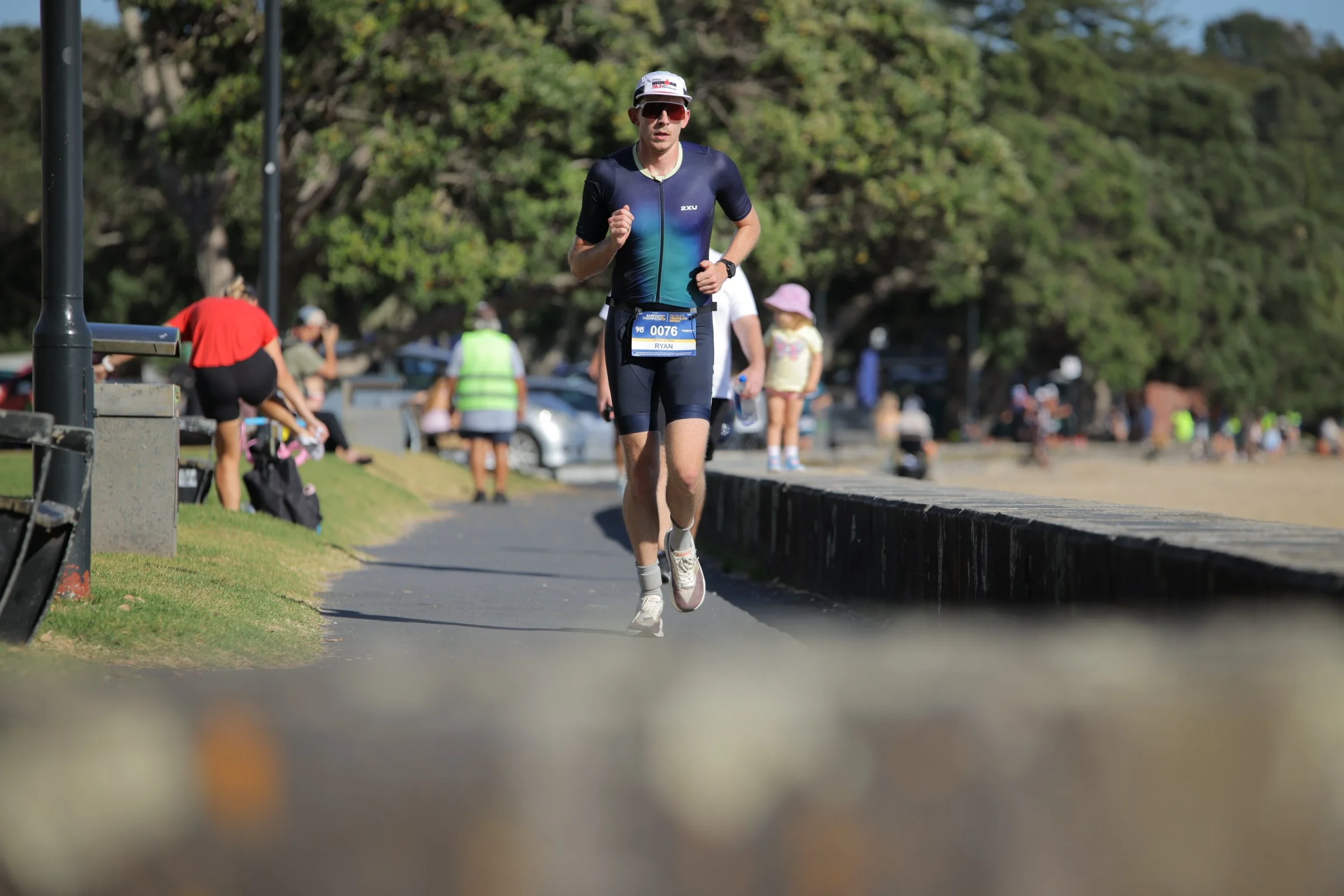 A man running on a paved path in a park, wearing athletic clothing, sunglasses, a cap, and a race bib with the name 'RYAN' and the number 0076, surrounded by other people and lush green trees.