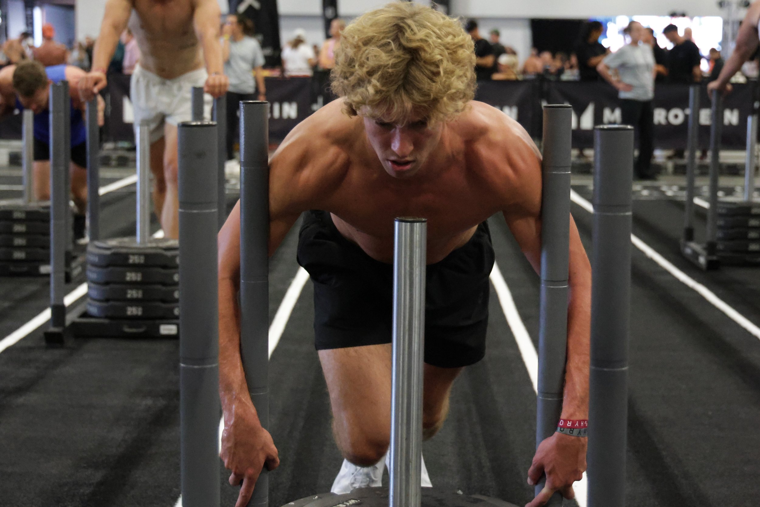 A shirtless man with curly blonde hair doing push-ups on a black gym floor with white lines, surrounded by other people in a busy workout gym.