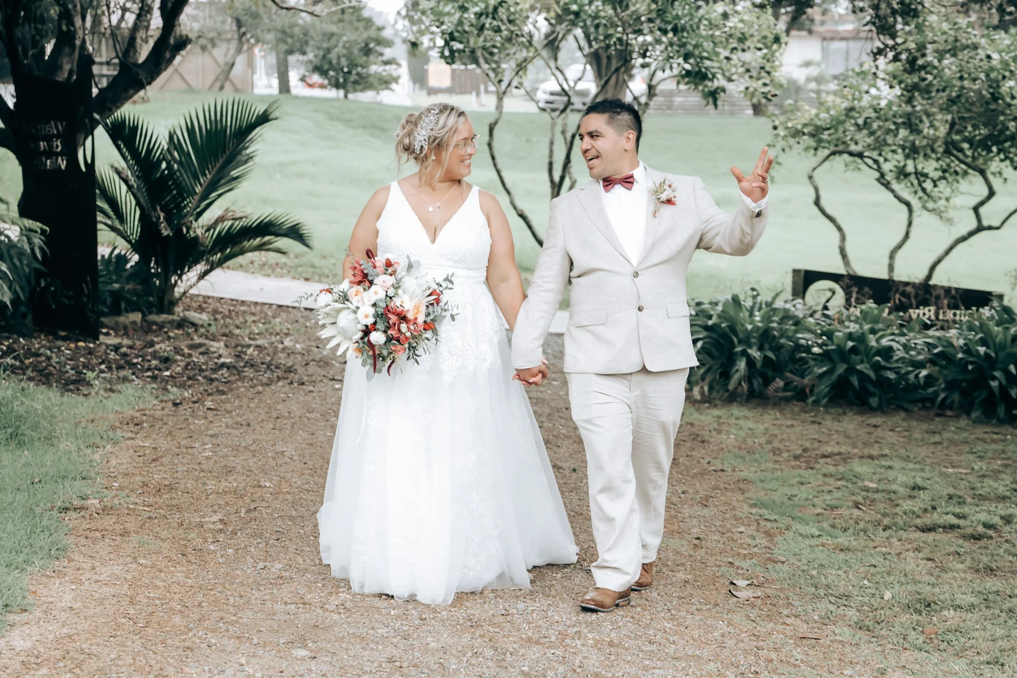 A bride and groom holding hands and smiling at each other while walking outdoors on a dirt path in a park with trees and greenery.