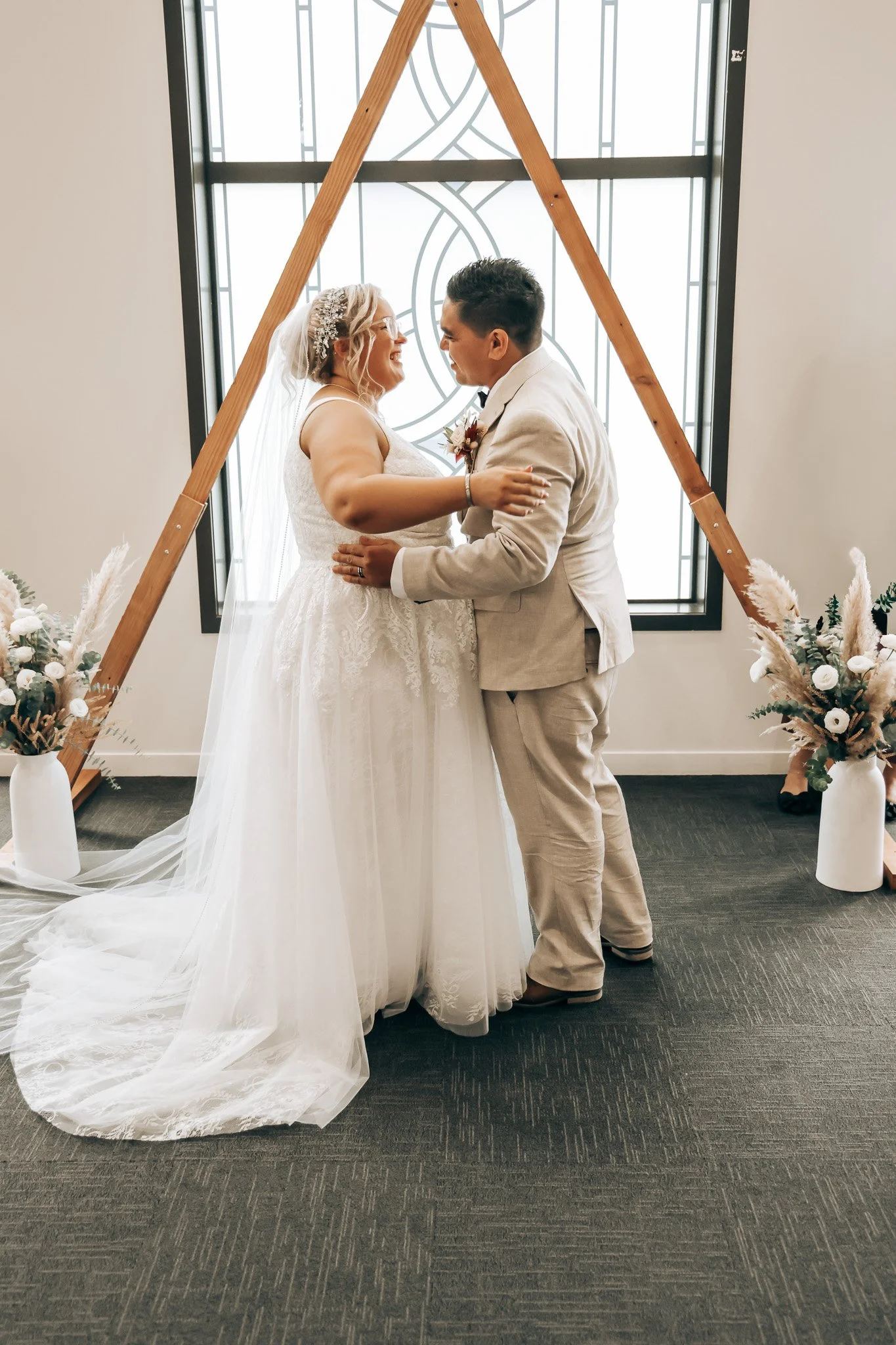 A bride and groom are standing close and smiling at each other during their wedding ceremony, inside a decorated venue with flowers and a large window with decorative ironwork in the background.