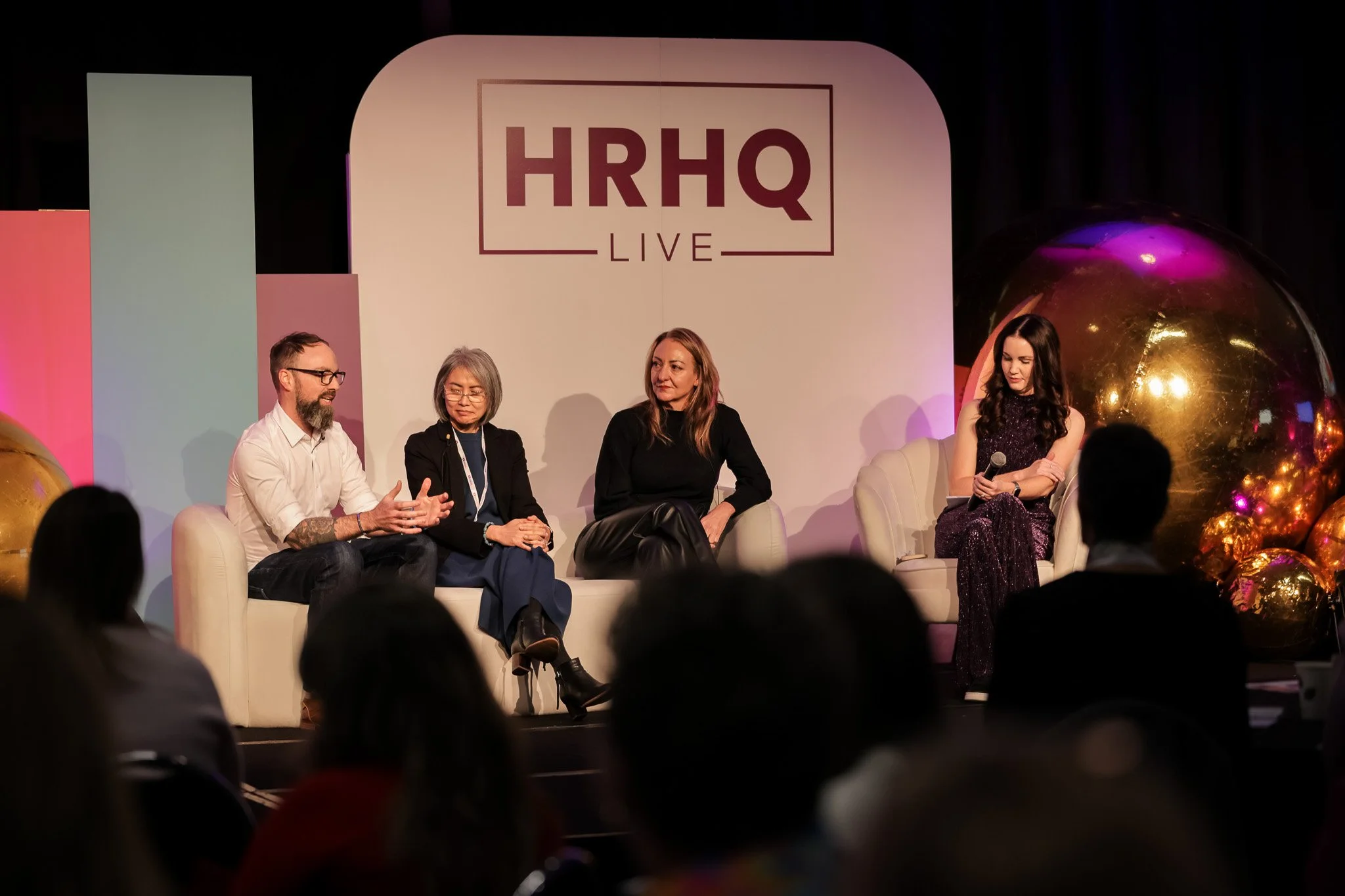 A panel discussion on stage with four people seated on a cream sofa and a woman on the right holding a microphone, with a large logo reading HRHQ Live in the background. Audience members are visible in the foreground, and there are decorative gold an