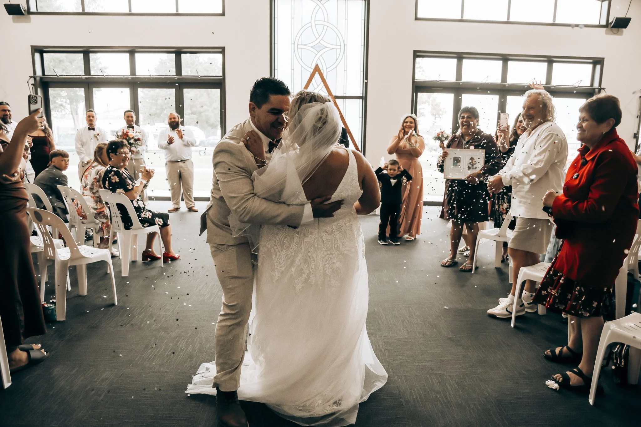 A bride and groom share a dance at their wedding reception, surrounded by guests seated and standing in a bright, modern venue with large windows. The guests are smiling and taking photos as confetti falls around them.