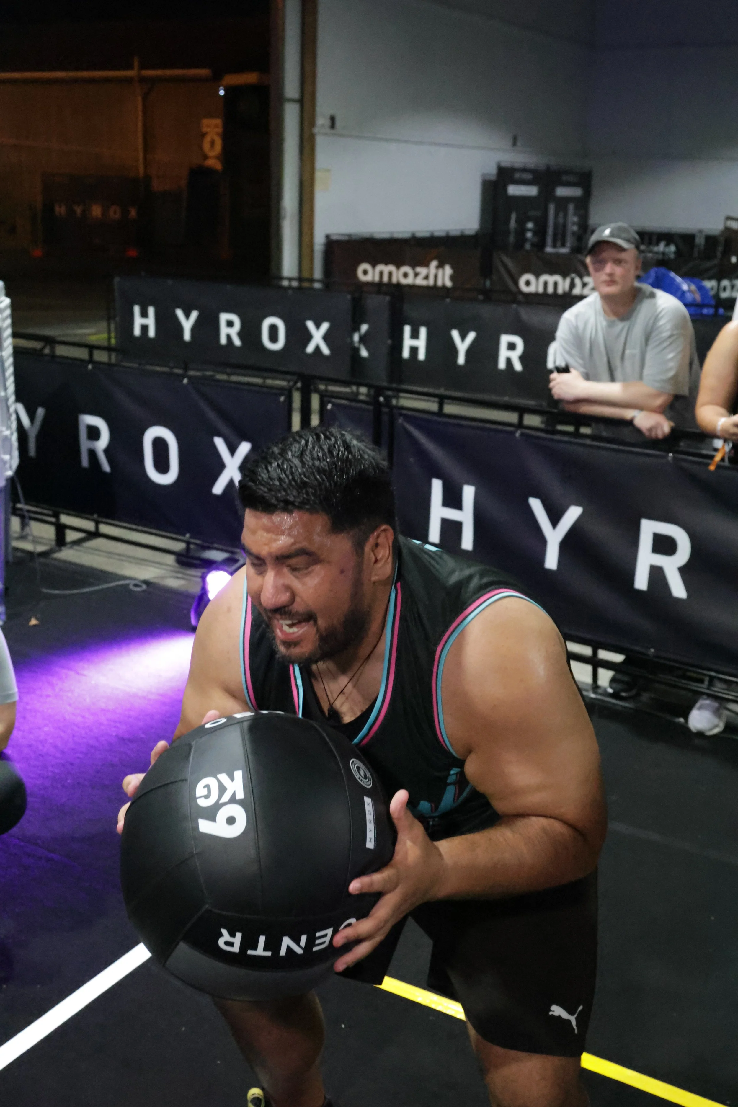 Man with dark hair and beard exercising with a medicine ball in a gym, seated on the floor with other people and gym equipment in the background.