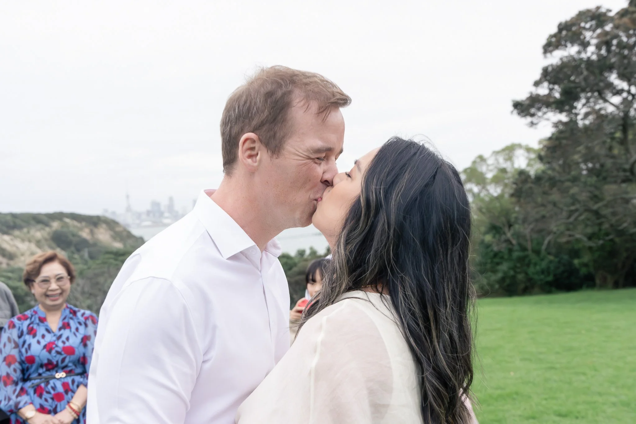 A couple sharing a kiss outdoors with a cityscape and green landscape in the background, while people watch and smile.