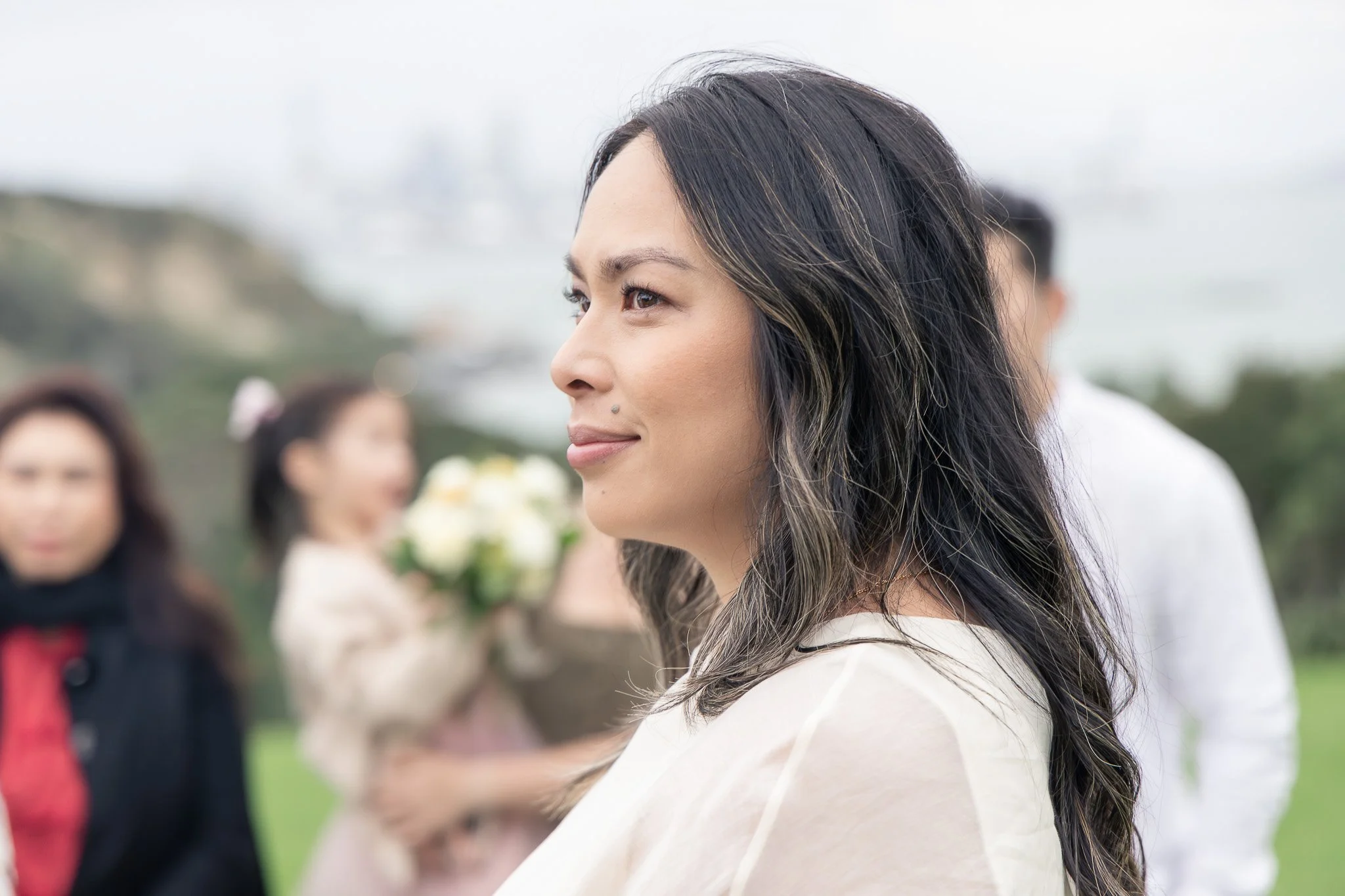 A woman with long dark hair and light highlights at an outdoor gathering, smiling softly, with blurred people in the background.