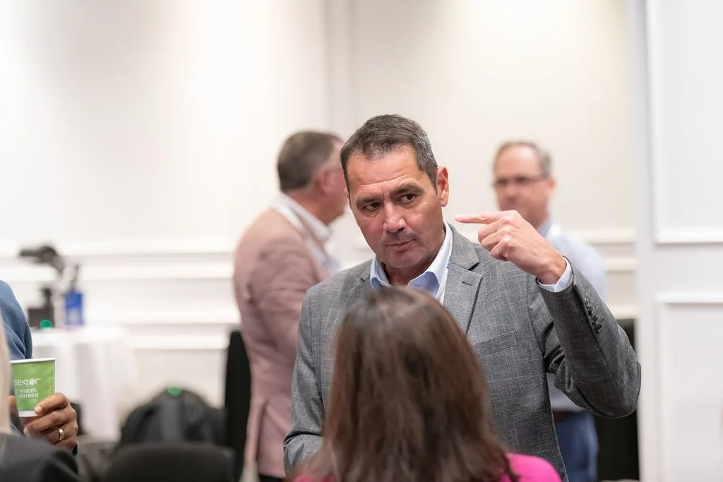 A man in a gray suit points to his head during a conversation at a conference or meeting, with other people in the background.