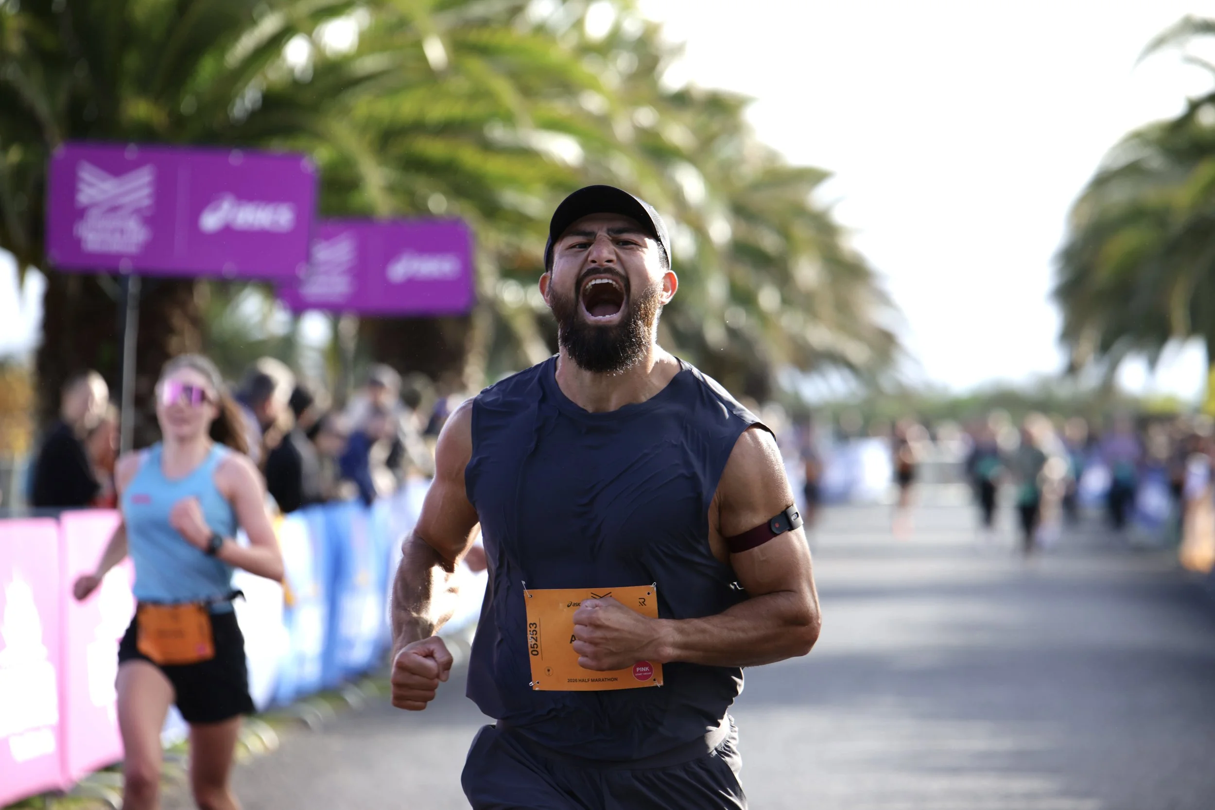 Male runner with a beard and dark cap, wearing a navy tank top, participating in a marathon, with many other runners and purple signs in the background.