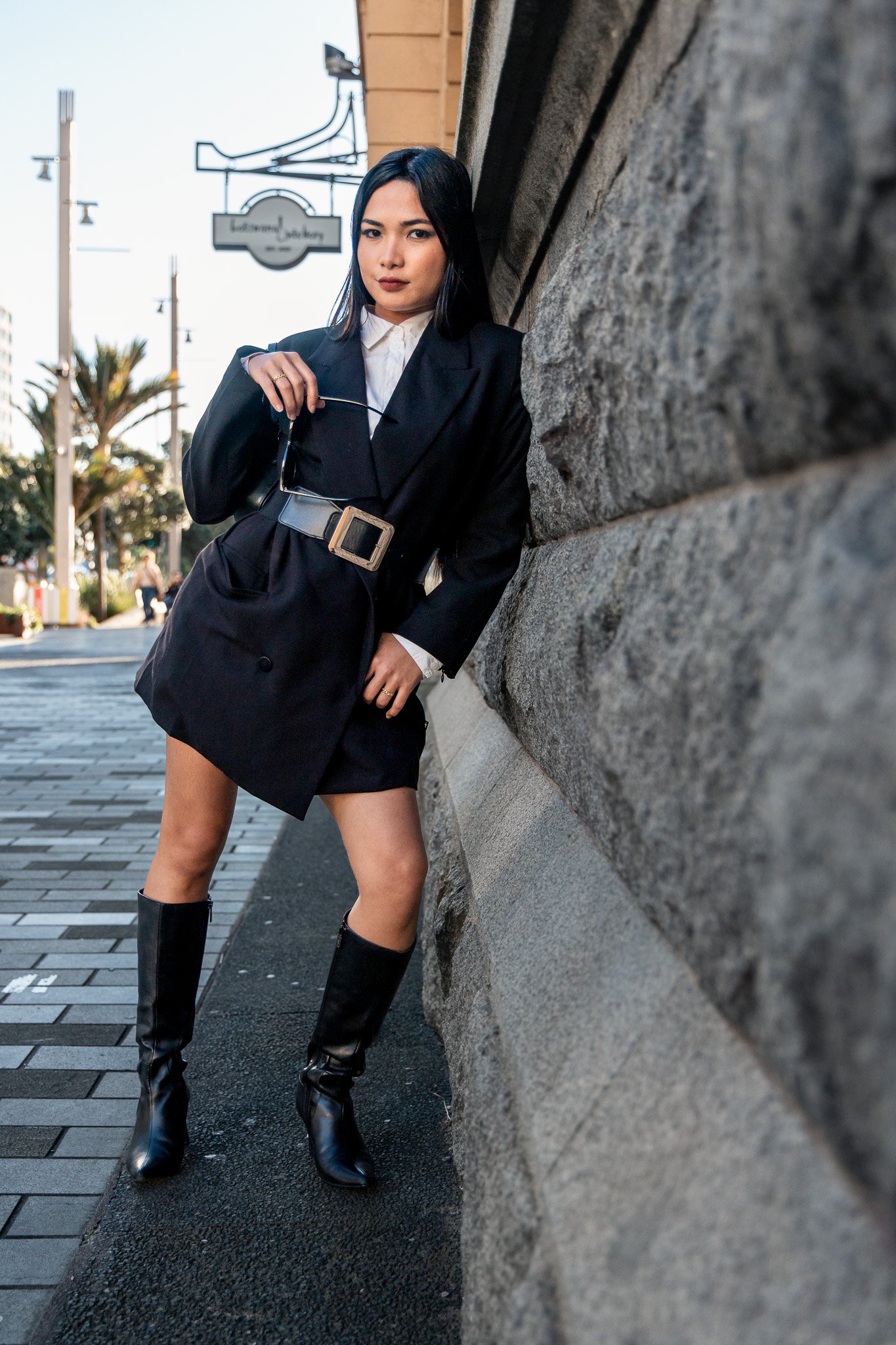 A woman with black hair and dark lipstick poses against a stone wall on a city street, wearing a black blazer-style dress, black knee-high boots, and holding sunglasses.
