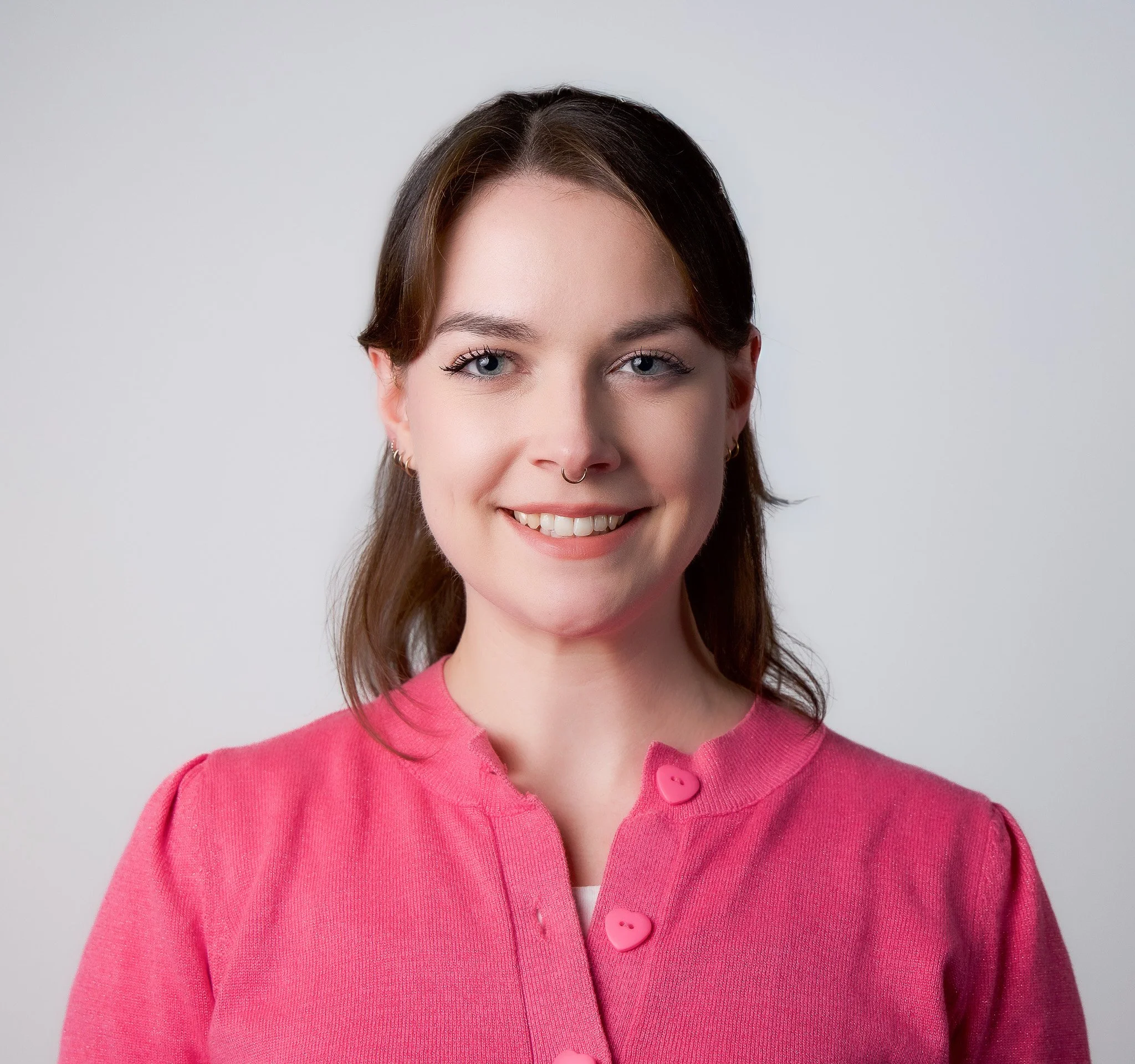 A woman with brown hair, blue eyes, and earrings wearing a pink cardigan with heart-shaped buttons smiling against a plain gray background.