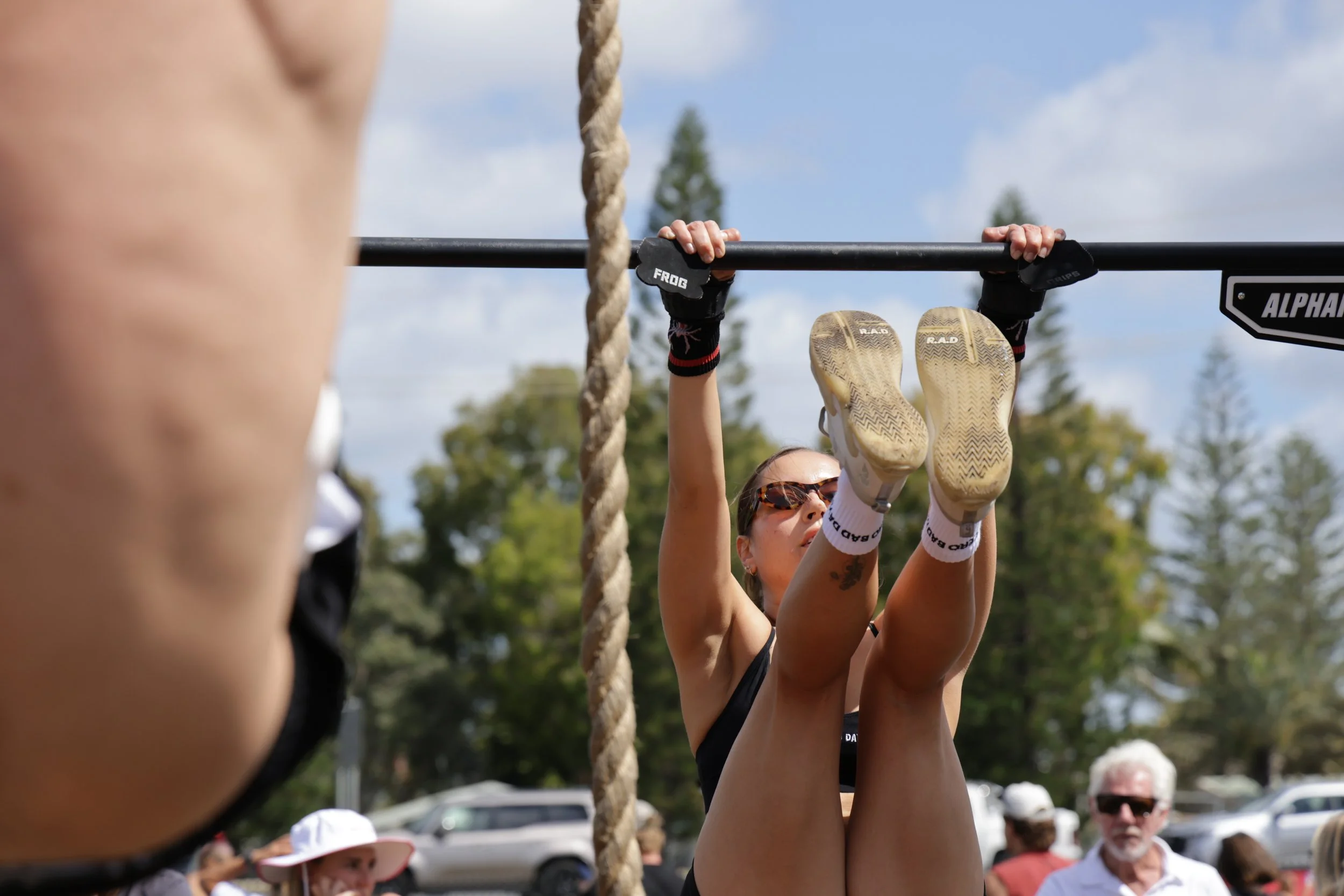 Participants participating in an outdoor event, hanging from gymnastic bars with a rope nearby, with a crowd of onlookers in the background.
