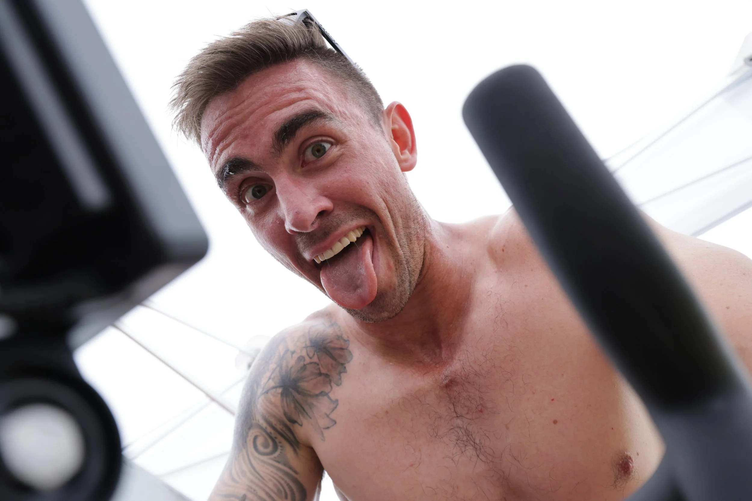 A shirtless man with tattoos on his arm and chest, smiling and sticking out his tongue, taken from a low angle through gym equipment.