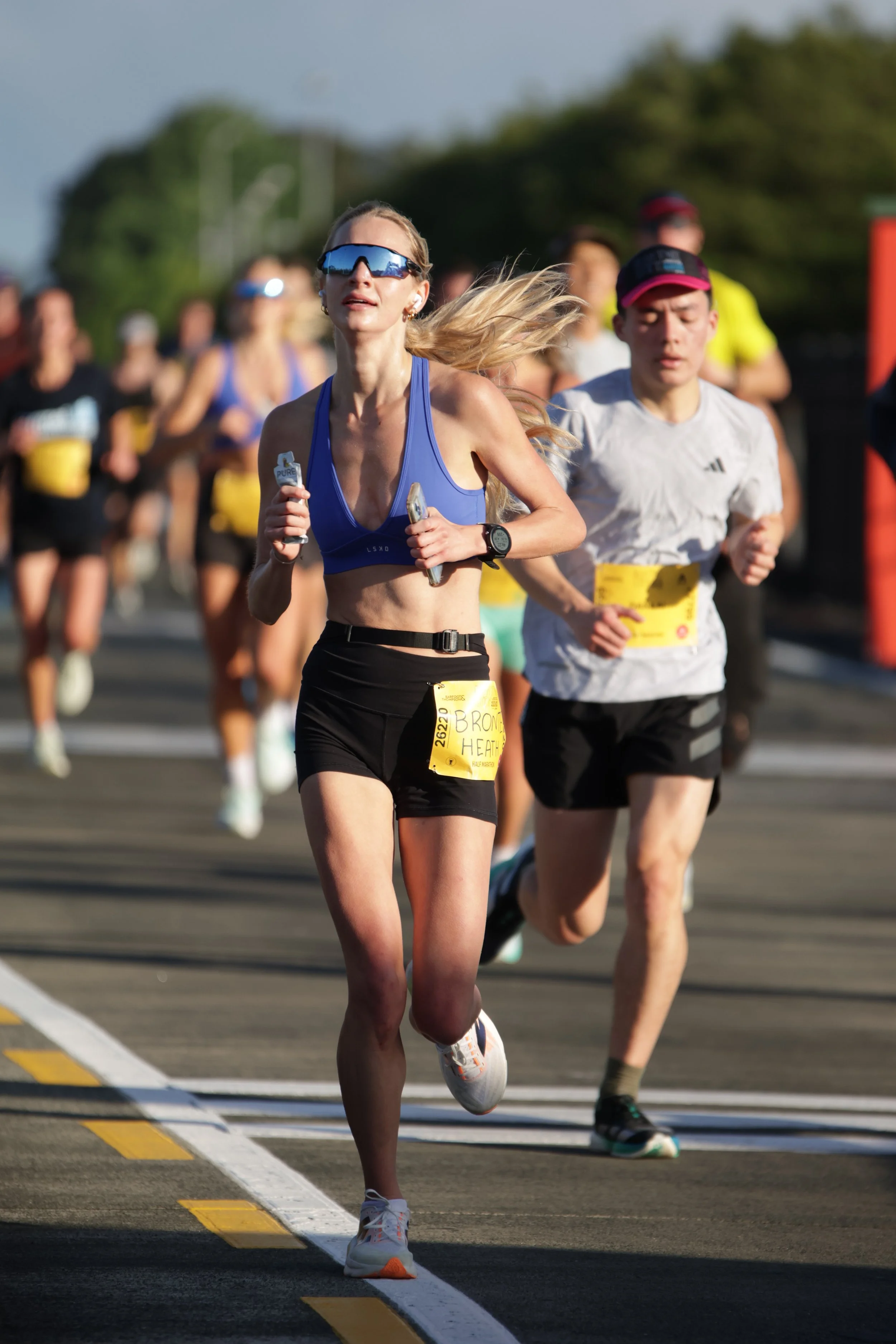 A group of marathon runners participating in a race. The woman in the foreground is wearing a blue sports bra, black shorts, sunglasses, and white running shoes. Other runners are visible in the background, running on a road with a yellow and white d
