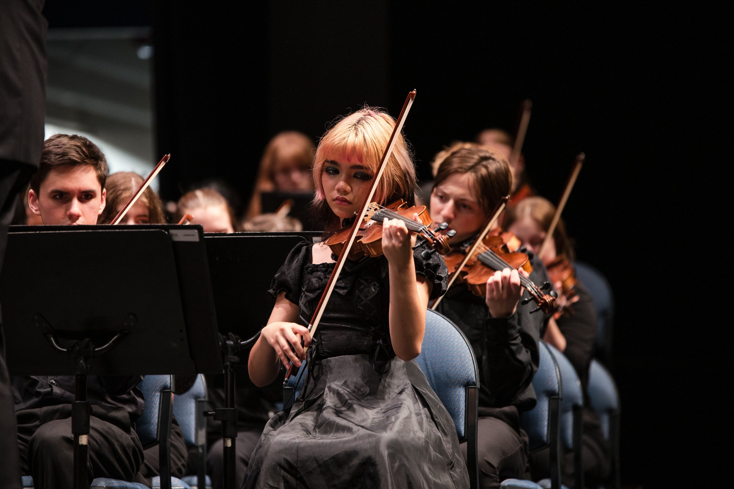 Young girl with pink hair playing a violin in an orchestra, surrounded by other musicians.