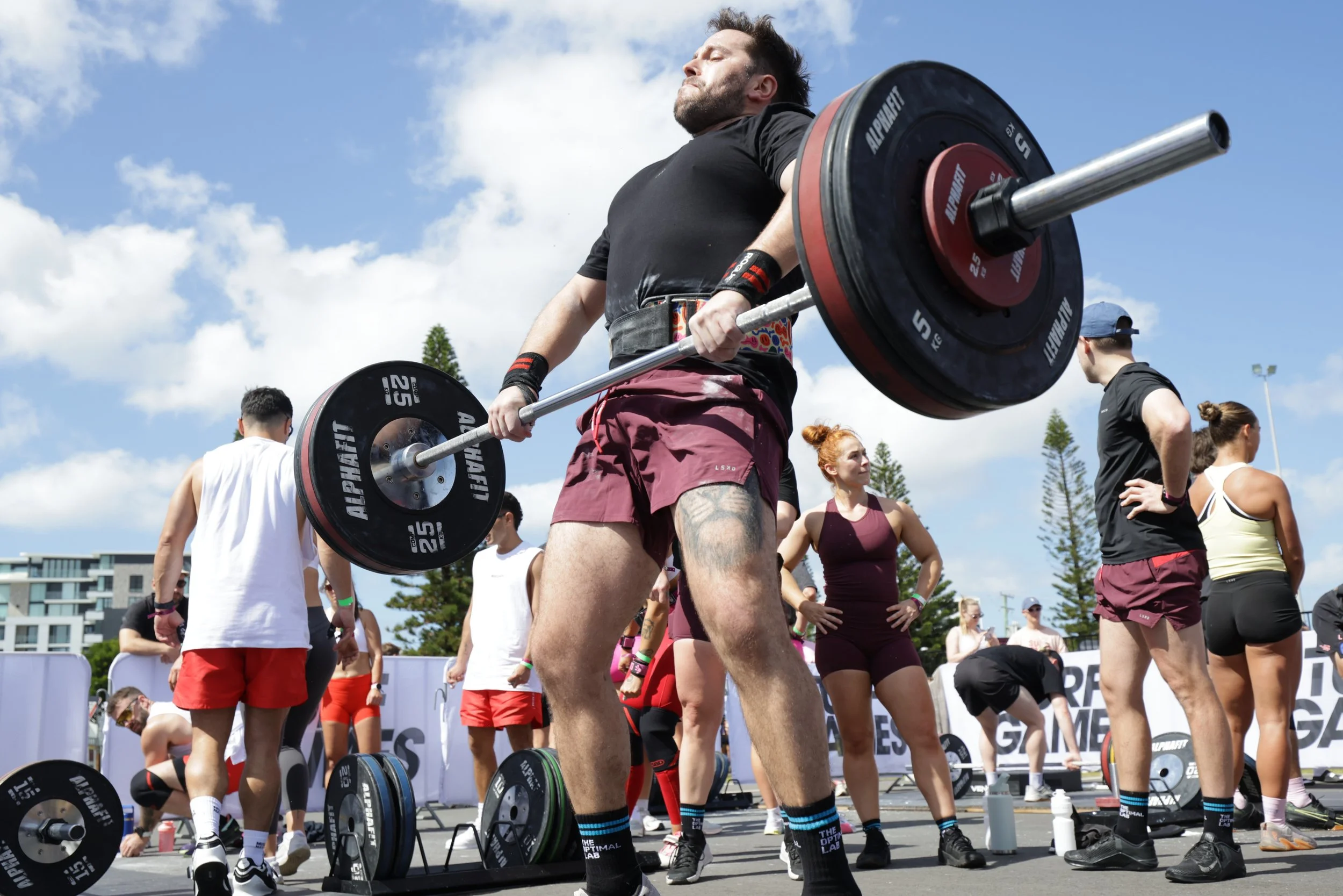Man lifting a barbell with weights at an outdoor fitness event, surrounded by other people in athletic attire.