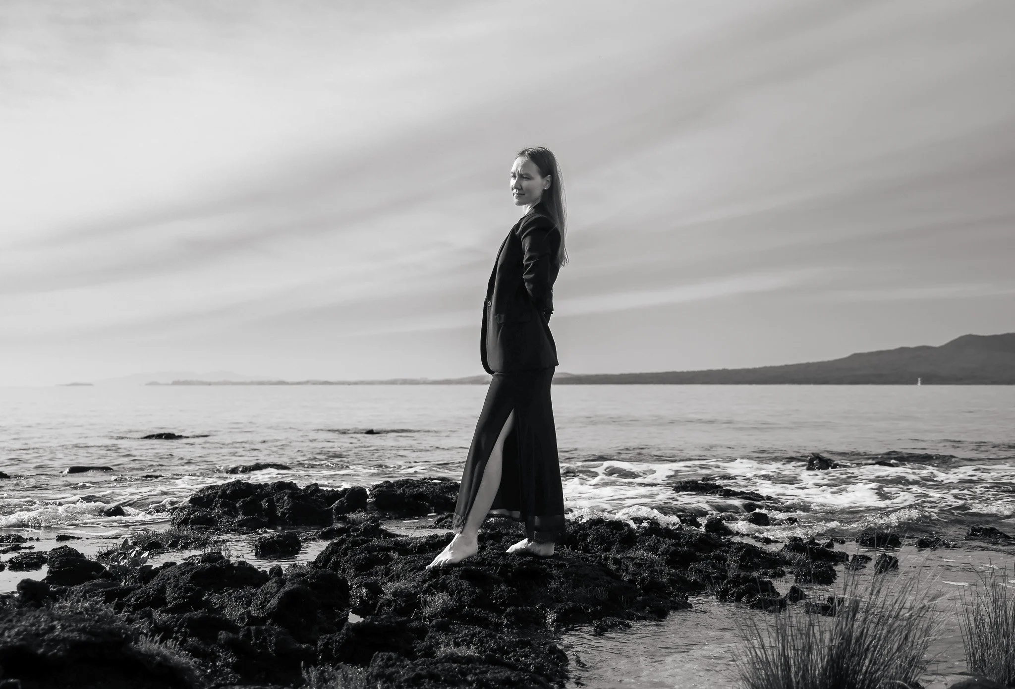 A woman in a black suit standing on rocks by the water at a beach during cloudy weather.