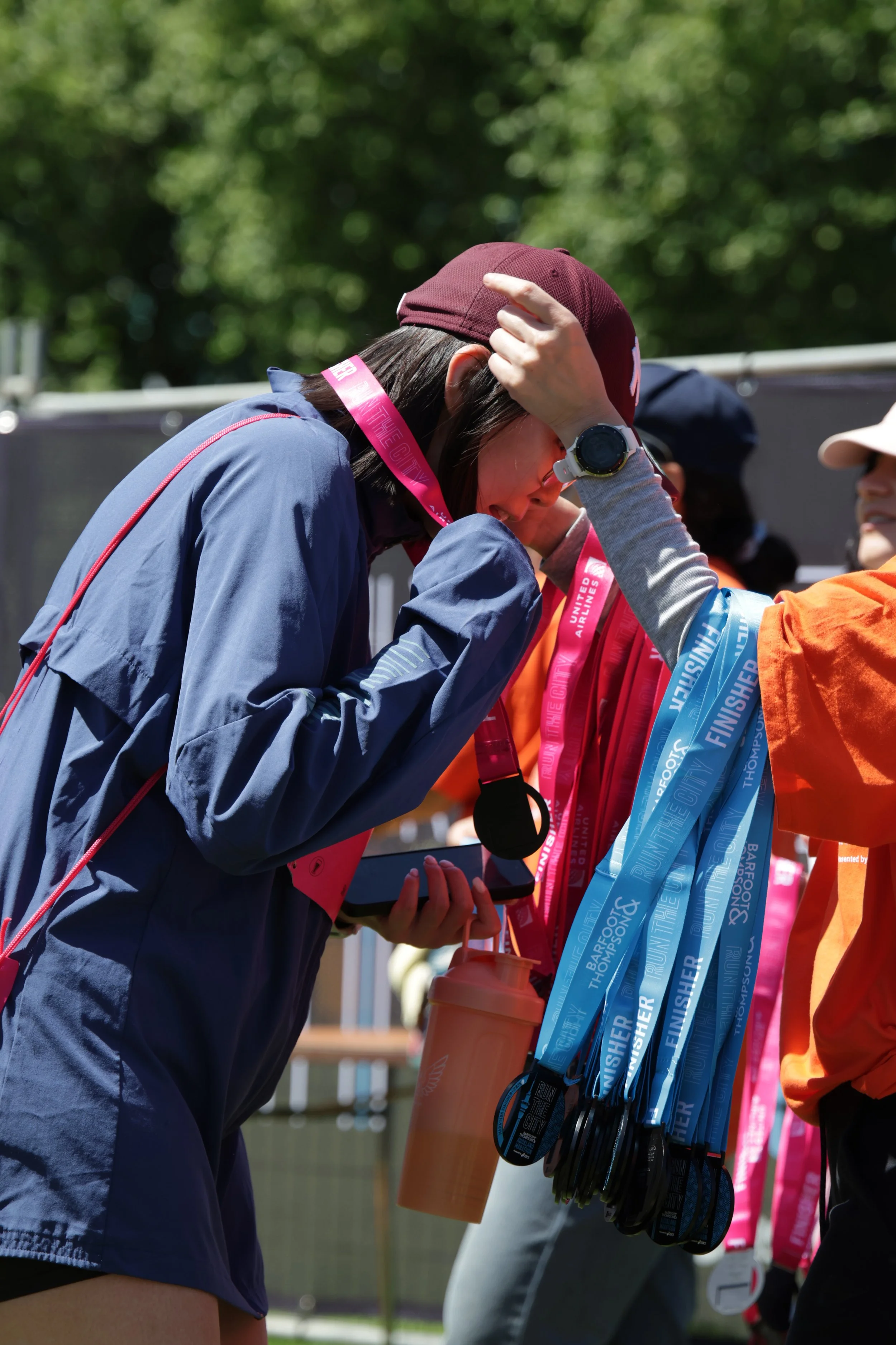 A person with a maroon cap and a wristwatch is receiving a finishing medal after a race, with blue medals hanging nearby. A participant holding a water bottle is partly visible in the background.