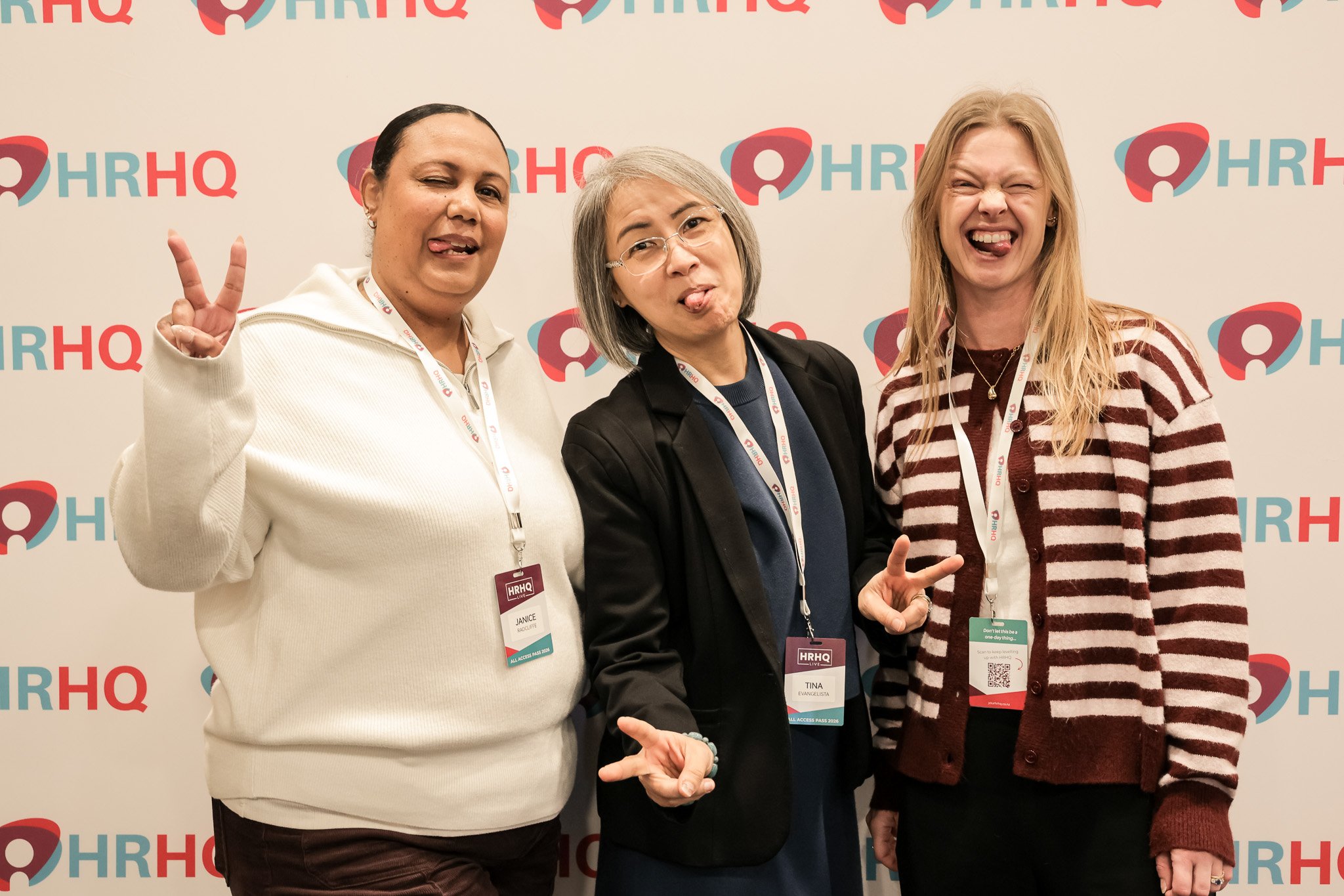 Three women standing in front of a HRHQ backdrop, making playful facial expressions and hand gestures at a professional event.