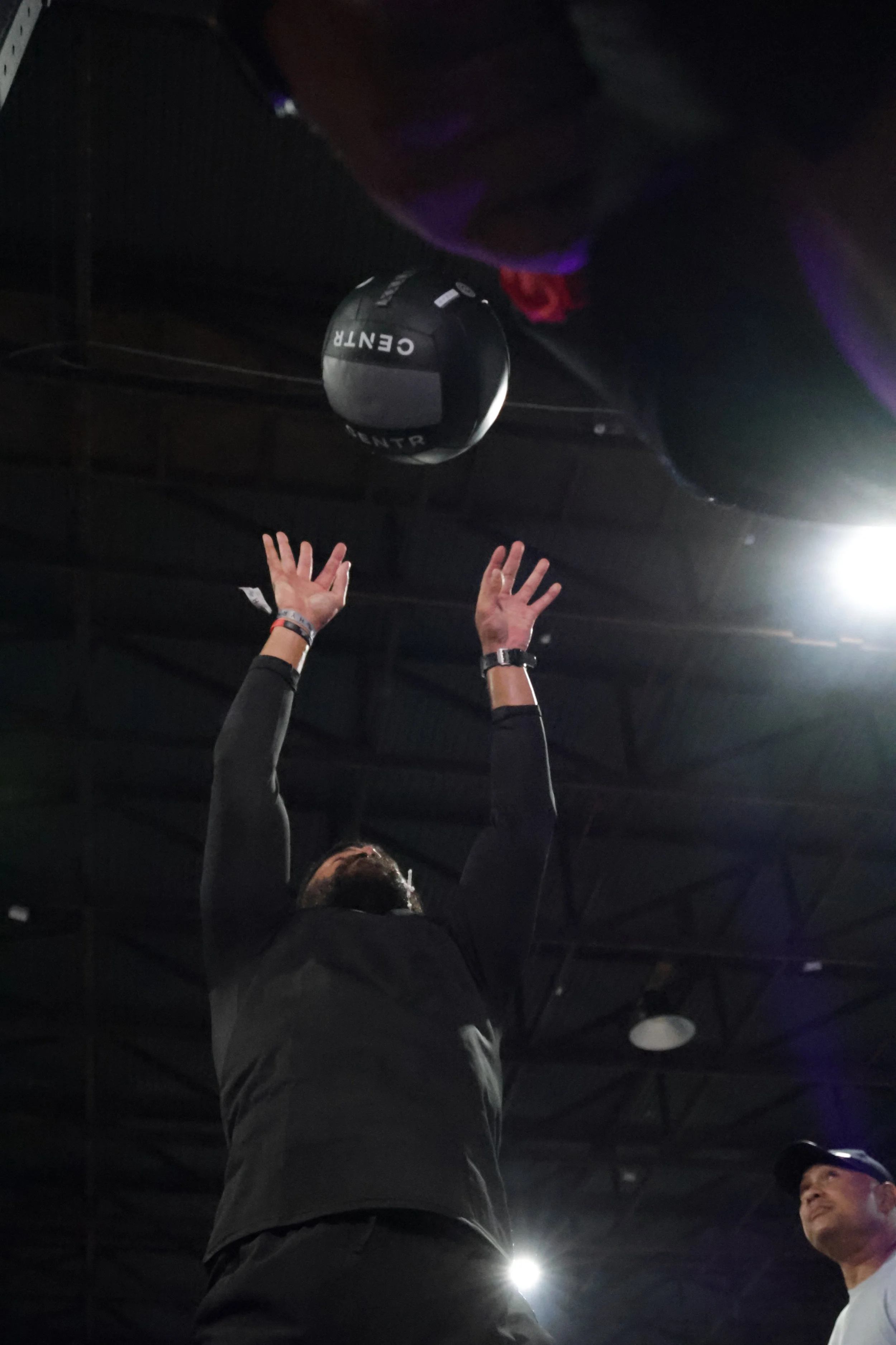 Person with black long sleeve shirt and wristbands, reaching up with both hands, in a dark indoor setting, with another person standing on the right side of the image, illuminated by overhead lighting.