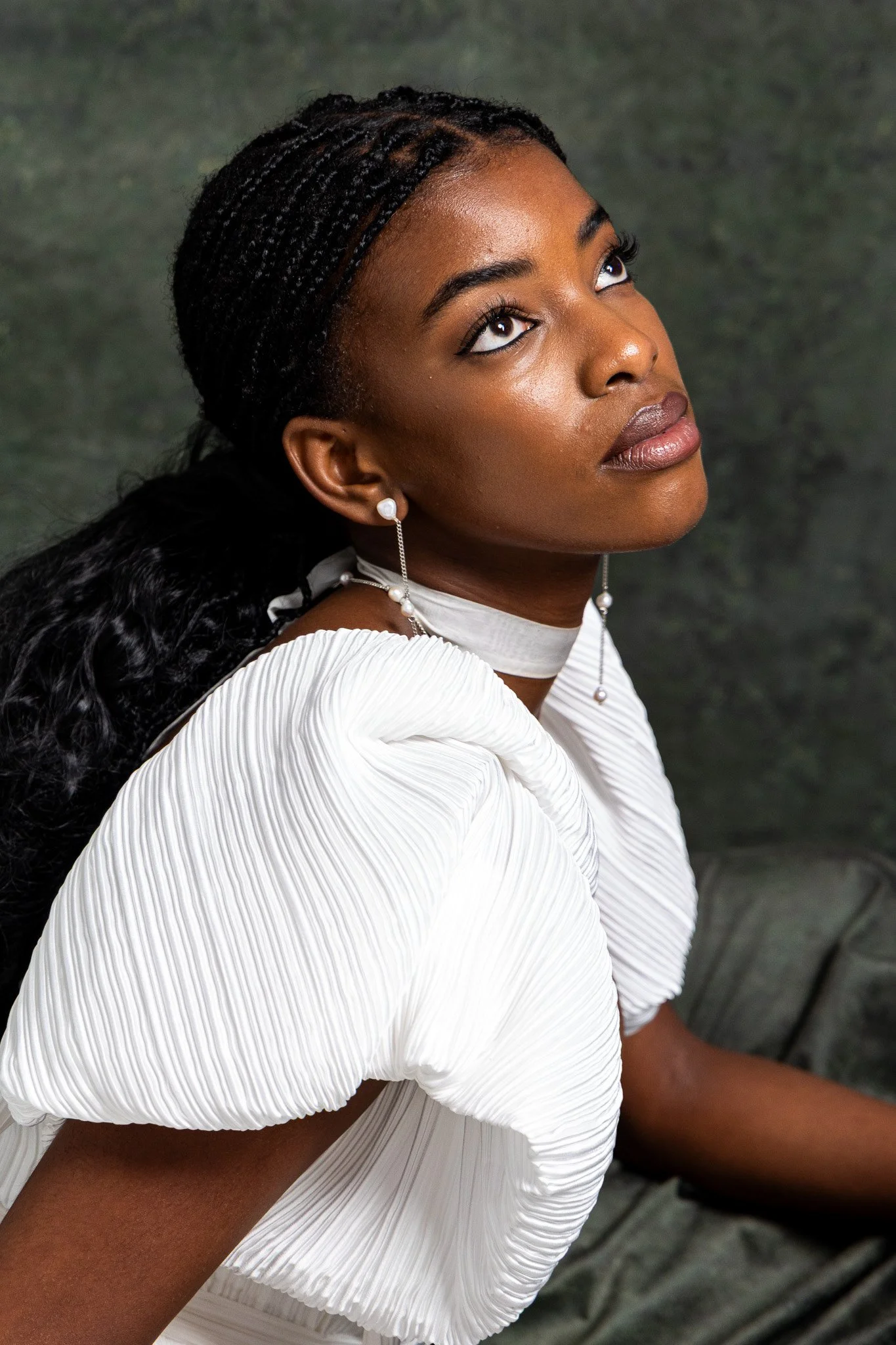 A woman with dark skin, black braided hair, and wearing a white pleated dress with puffed short sleeves, looking upwards. She has pearl earrings and a pearl necklace, and the background is dark and textured.