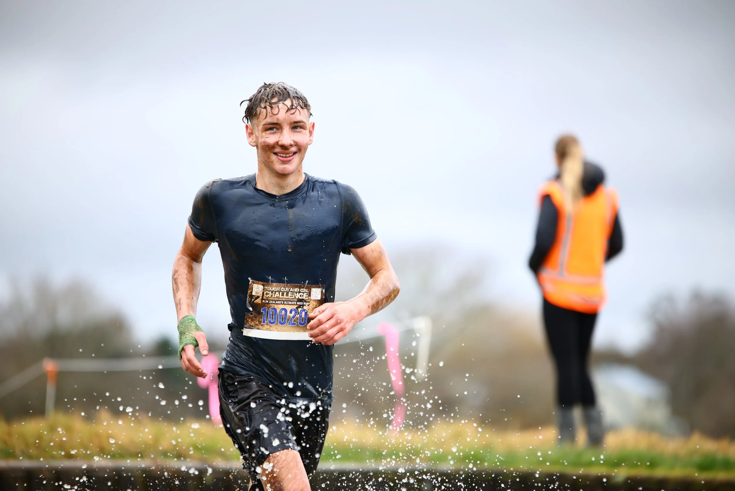 Young man running through water during an outdoor challenge race, smiling, with a female volunteer in the background