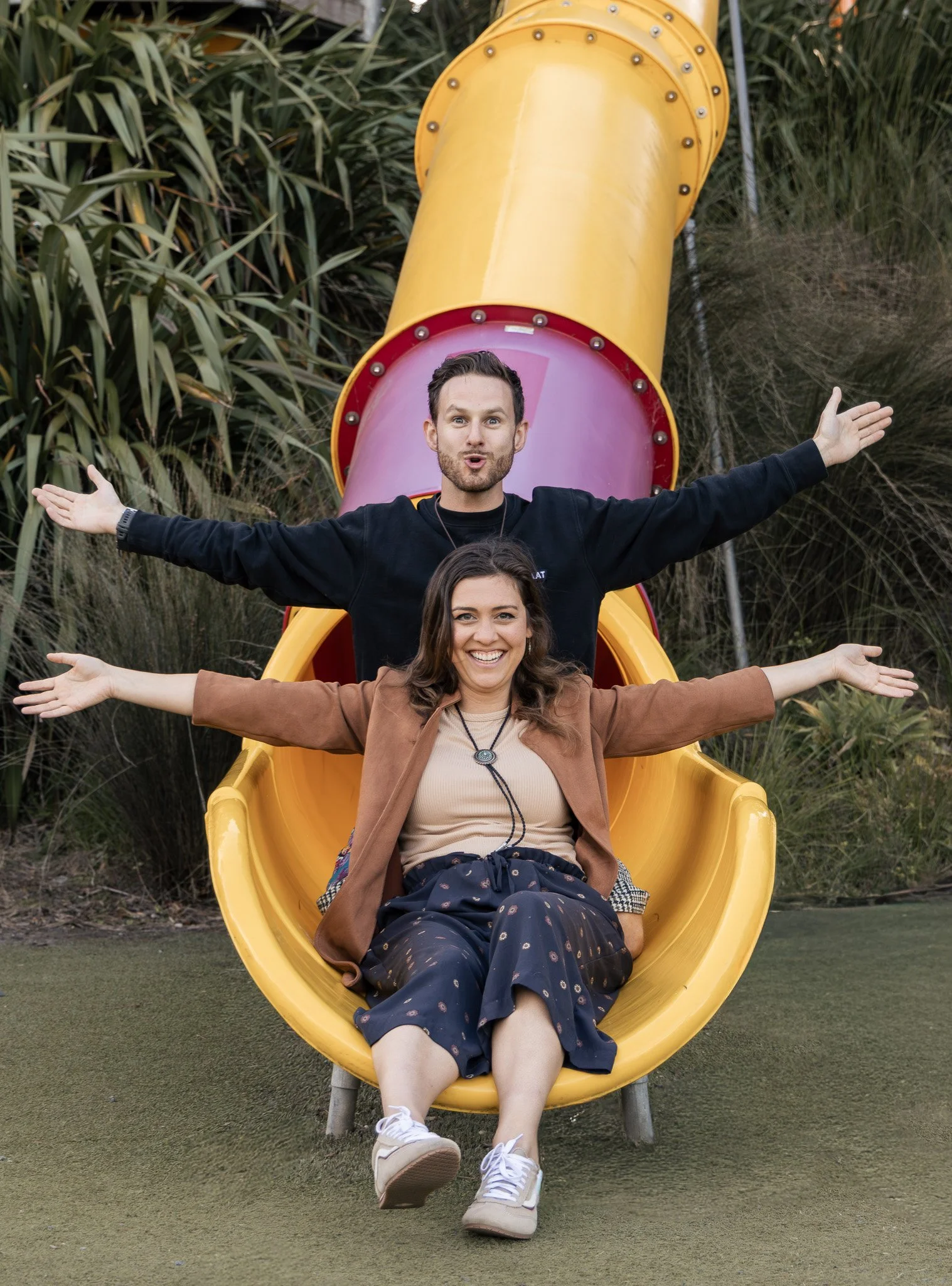 Two women and one man sitting at the top of a yellow slide at a playground. The man is on top with arms outstretched, and the women are sitting below him, smiling with arms extended to the sides. They are outdoors with grass and bushes in the backgro