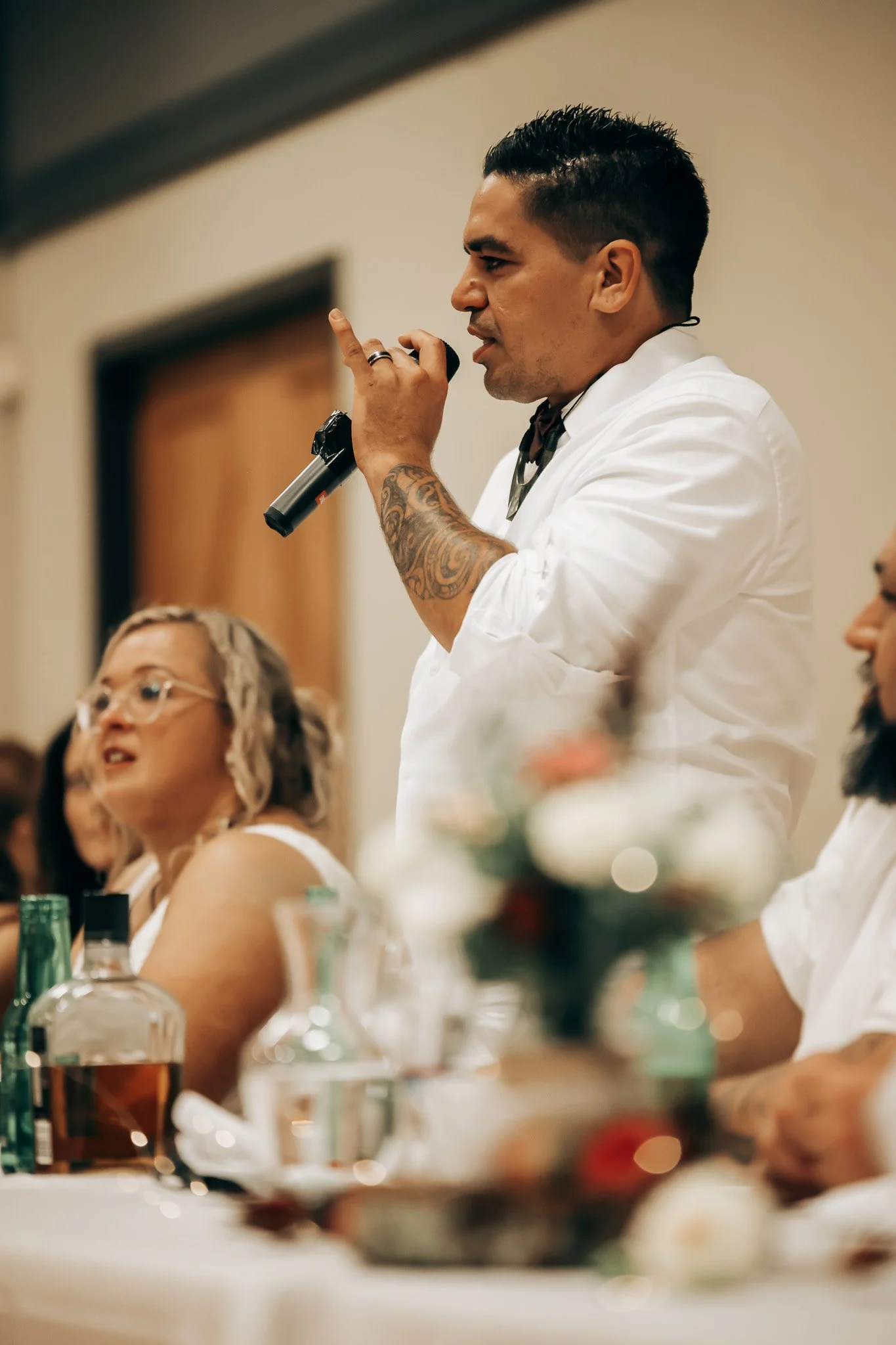 A man with dark hair, tattoos on his arm, and wearing a white shirt is speaking into a microphone during a formal event. He is seated at a long table with other people, with bottles and glasses in front of him. The background shows a beige wall and a