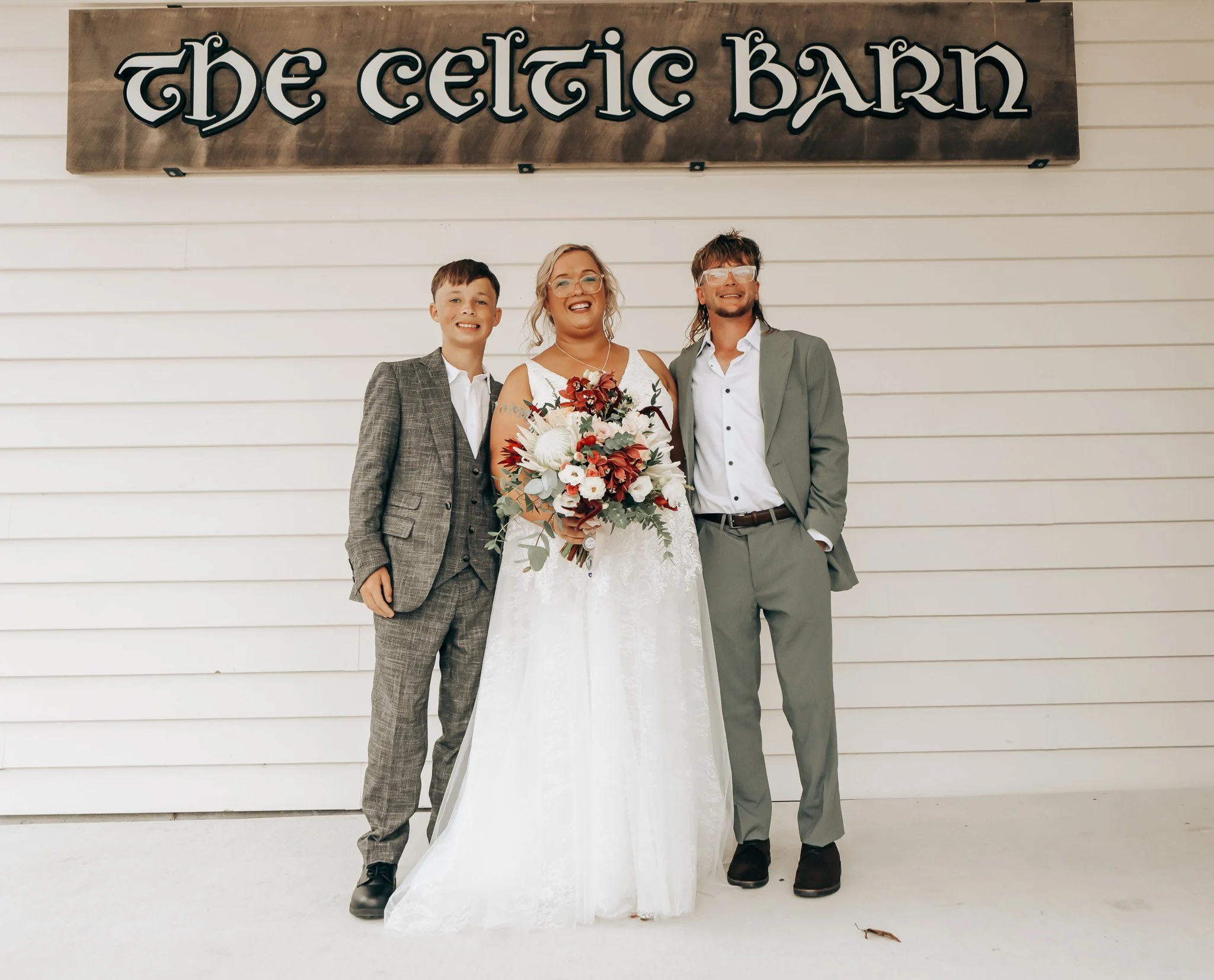 A bride in a white wedding dress holding a bouquet, flanked by two men in suits, standing in front of a sign that reads 'The Celtic Barn.'