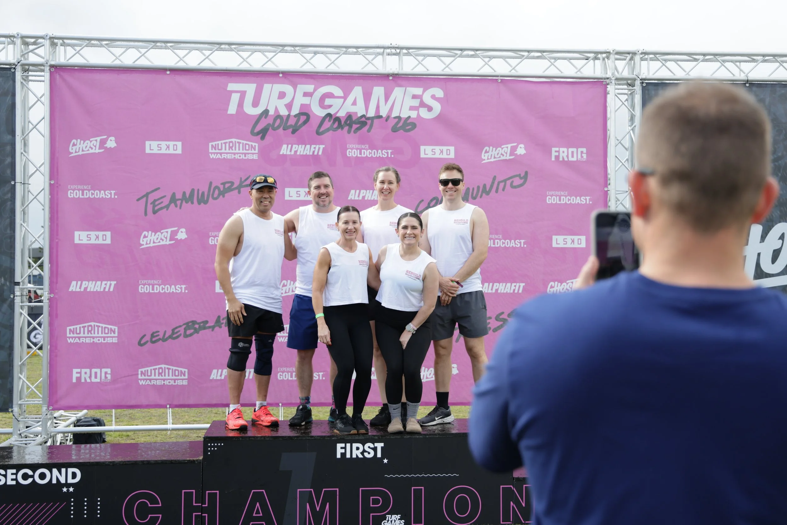 A group of six people, four women and two men, posing on a winners' podium at an outdoor event with a pink backdrop. One person is taking a photo of them with a smartphone. The backdrop features various sponsor logos and event branding for "TURF GAME