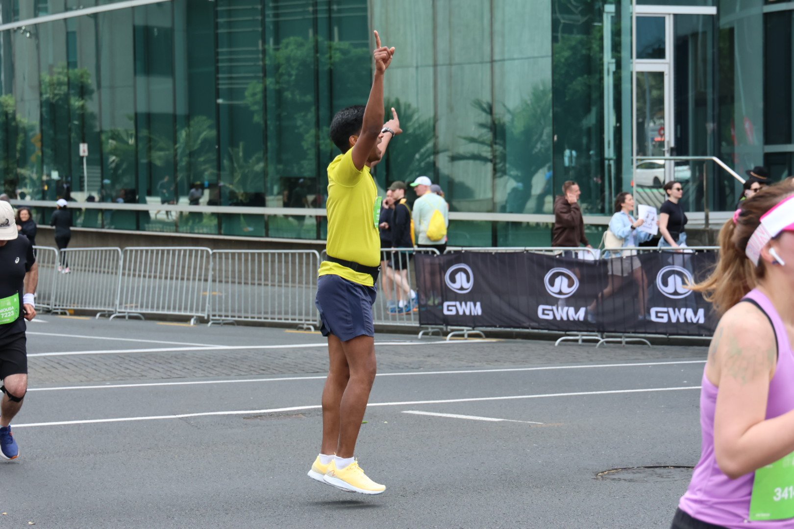A man in a yellow shirt and shorts jumping in the air during a sports event or marathon, with other participants and spectators in the background.