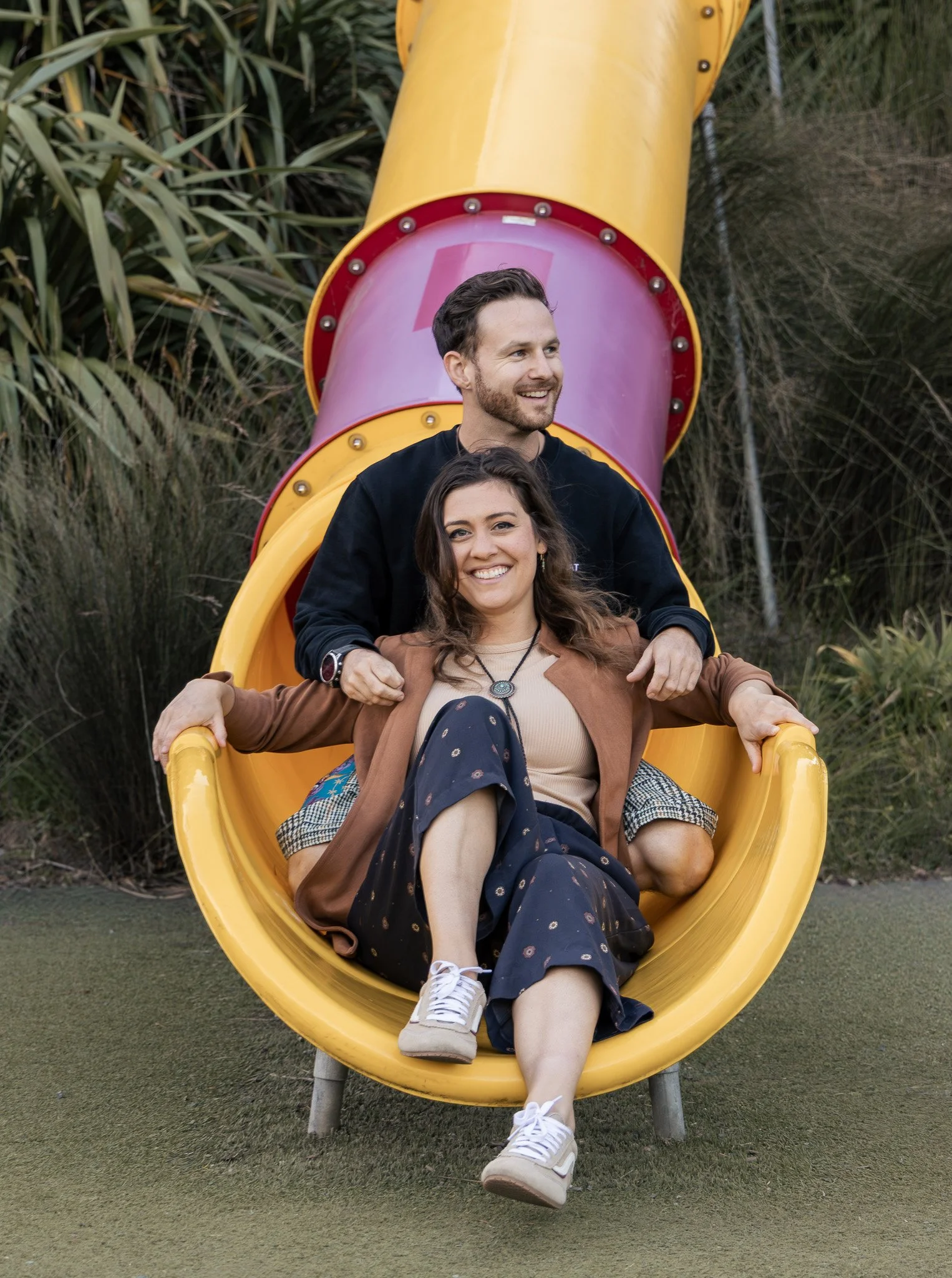 Two smiling people, a man and a woman, sitting inside a yellow playground slide in an outdoor park area surrounded by green plants and grass.