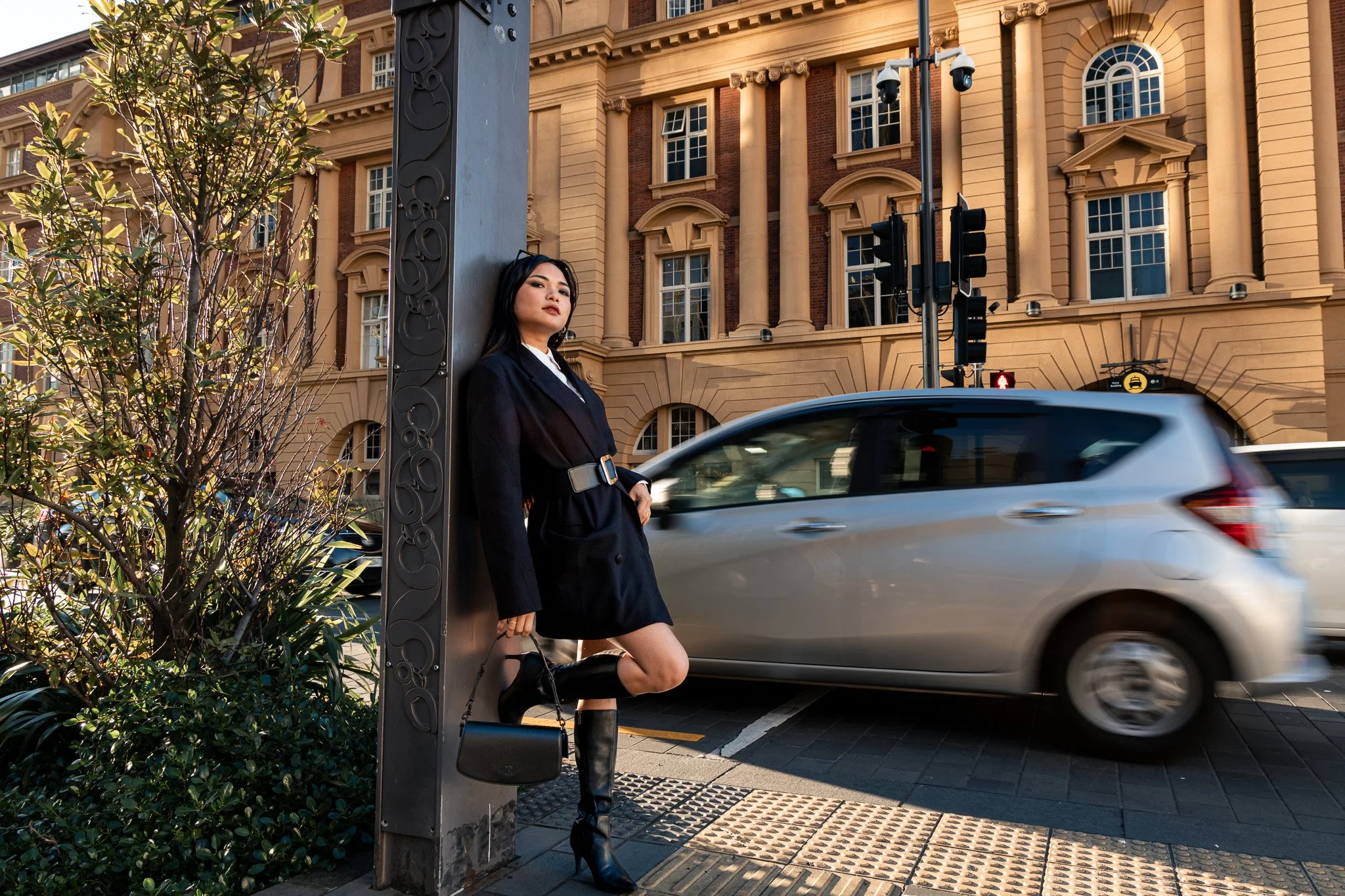 A woman dressed in a black blazer and skirt, with black knee-high boots, standing on a city sidewalk near a crosswalk, leaning against a metal pole near a traffic light. She is holding a small black purse. There is a tree with green leaves and bushes