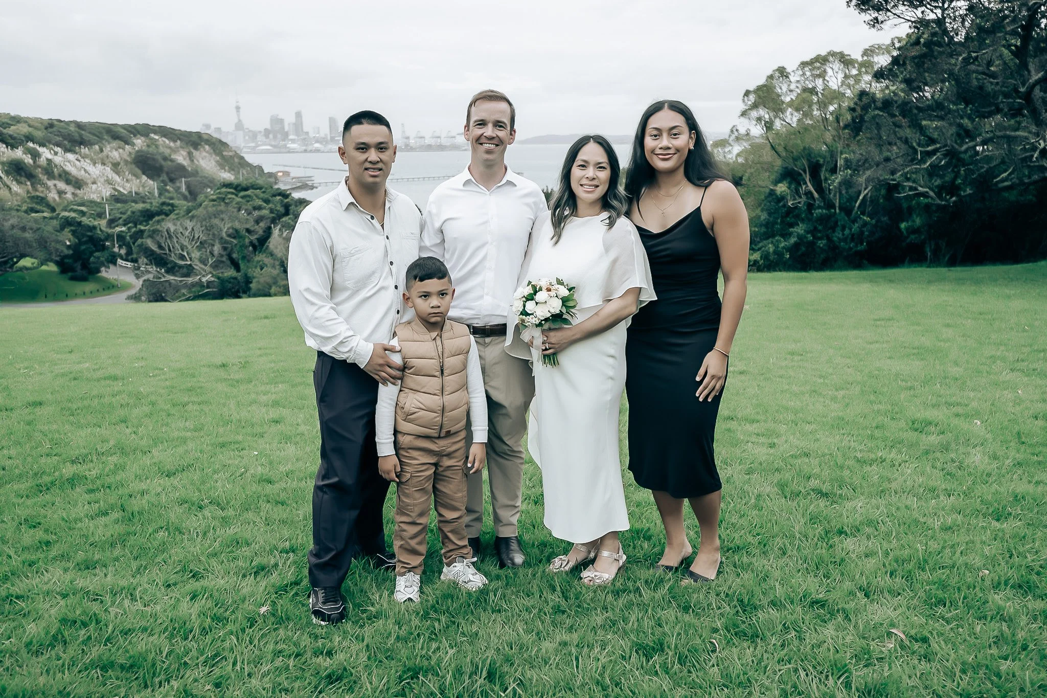 Group of five people standing outdoors on a green lawn with city skyline and water in the background, dressed in formal and casual clothing, smiling at the camera.