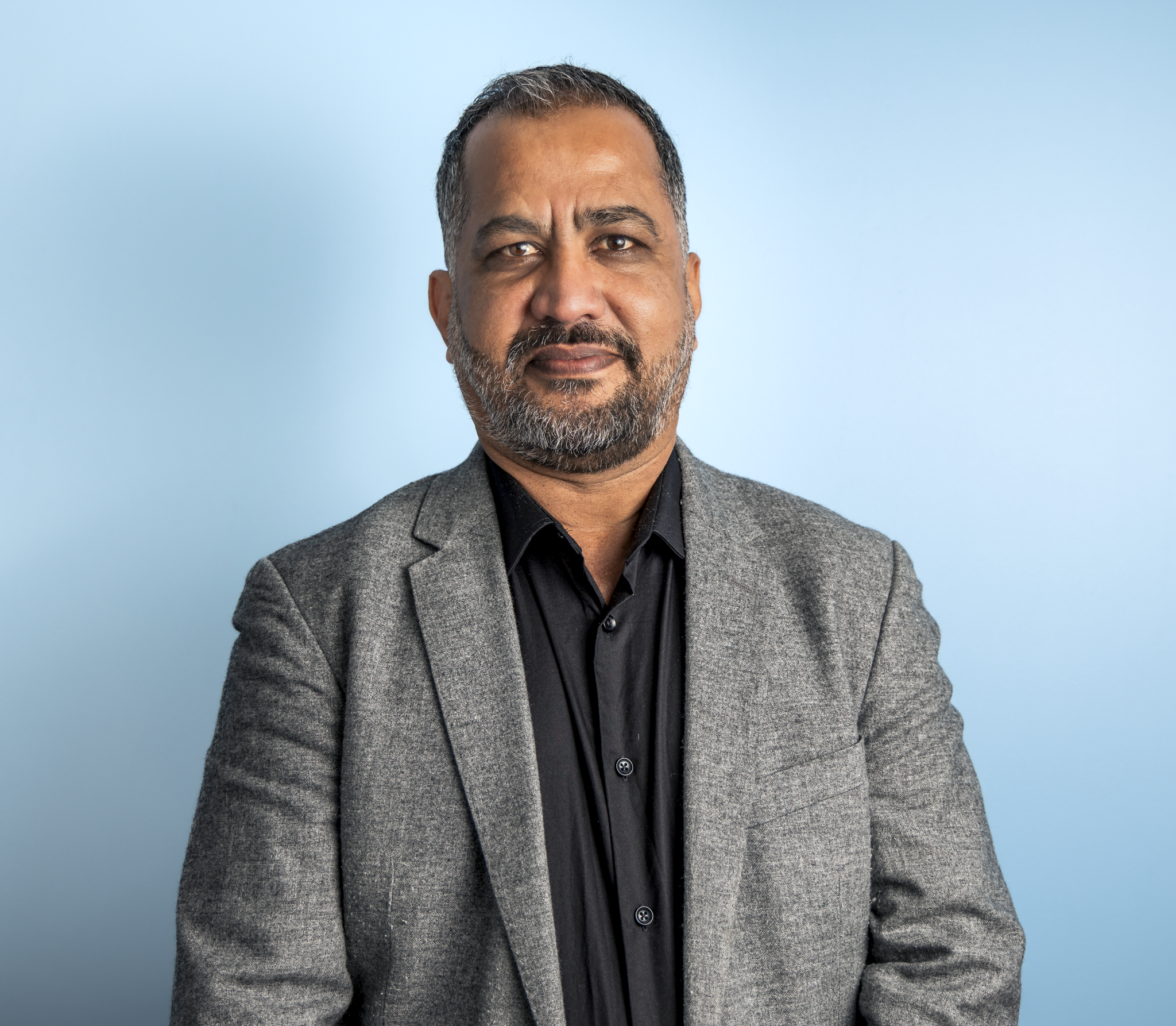A middle-aged man with short, dark hair and a beard, dressed in a gray blazer and black shirt, posing against a light blue background.