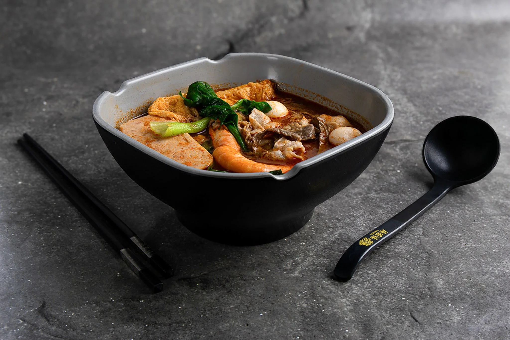 A bowl of ramen with vegetables and eggs on a dark surface, accompanied by black chopsticks and a black spoon.