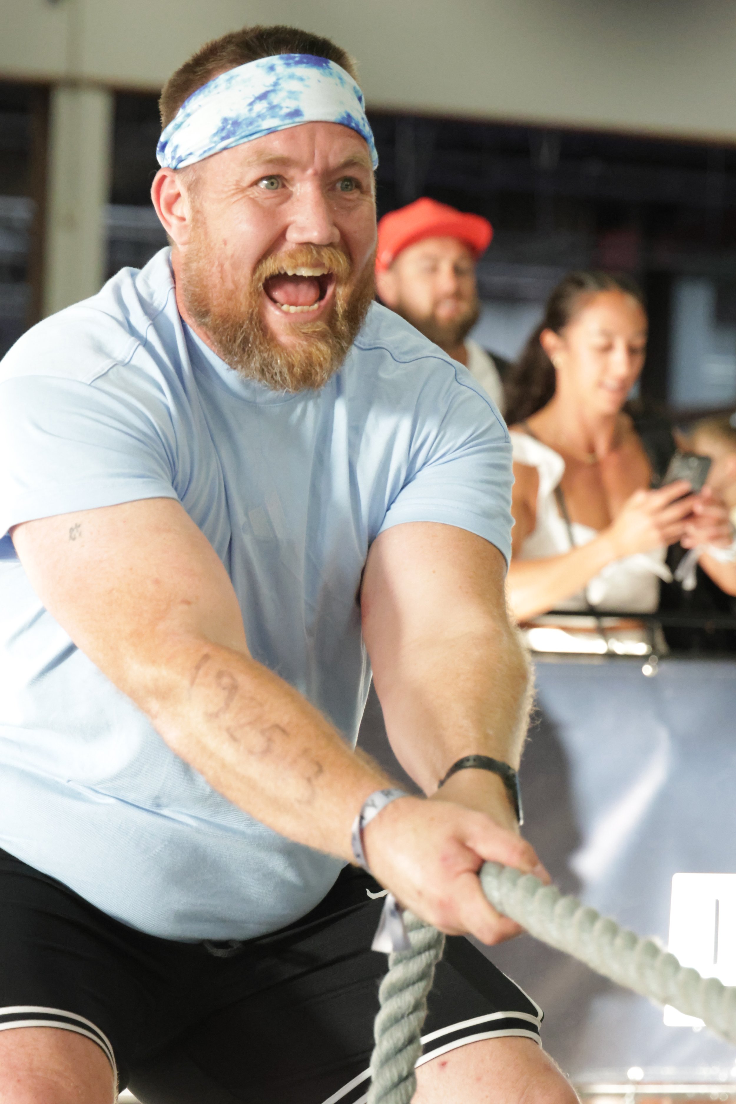 A man wearing a blue and white tie-dye headband and a light gray T-shirt, with a beard and mustache, is passionately cheering or shouting during an indoor event, with two women in the background, one wearing a red hat and the other looking at her pho