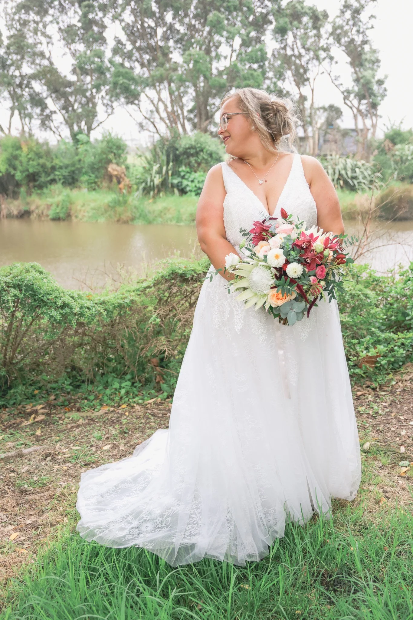 A bride in a white wedding dress holding a bouquet of flowers, standing outdoors near a river and green bushes, with trees in the background.
